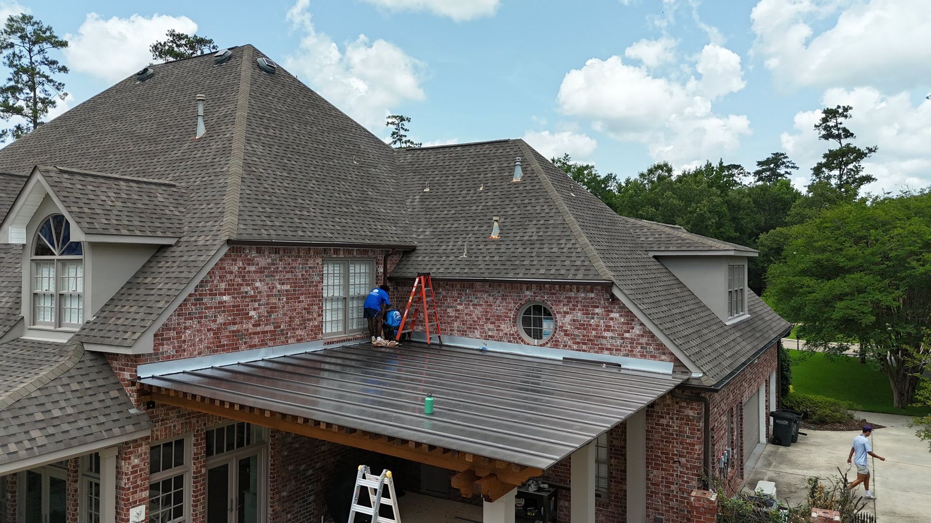 Workers install shingles on a patio roof attached to a brick house on a sunny day.