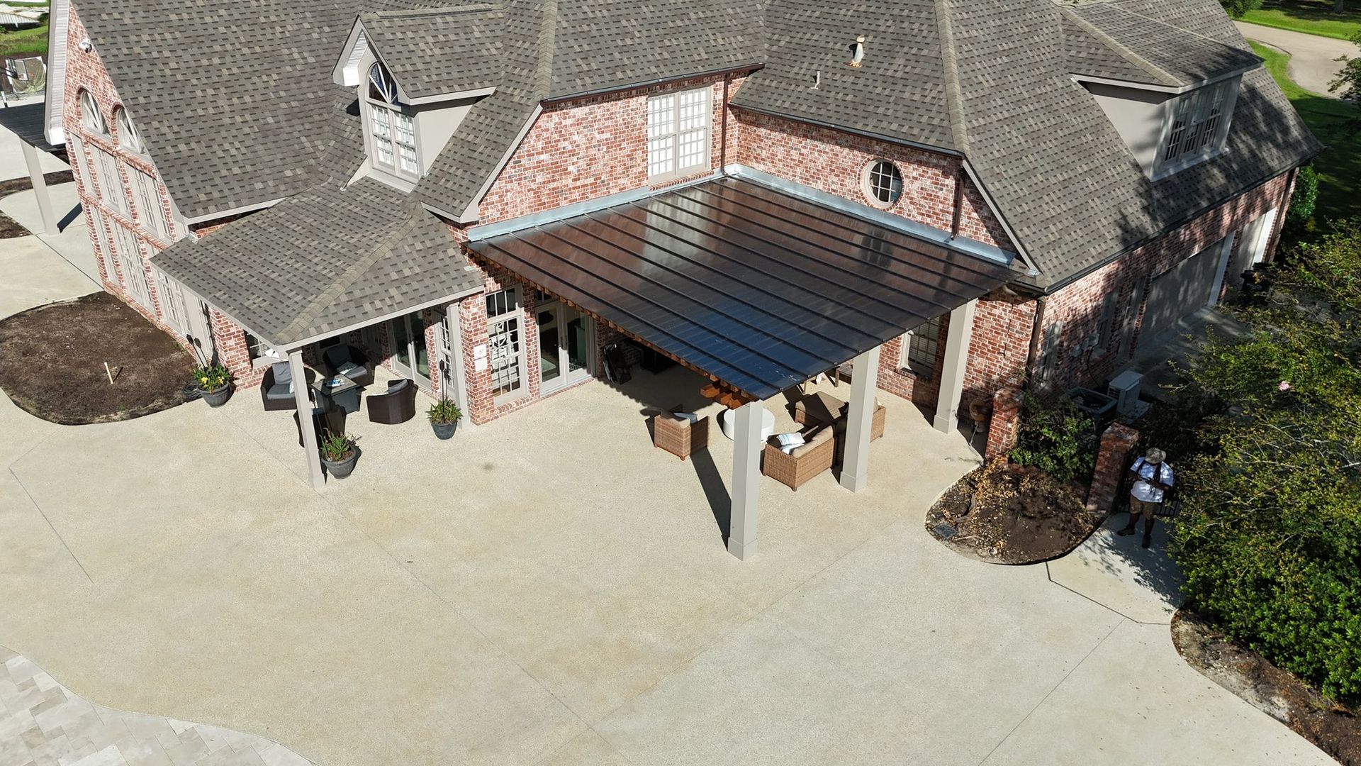 Aerial view of a brick house with a dark metal patio cover, outdoor furniture, and a concrete patio.