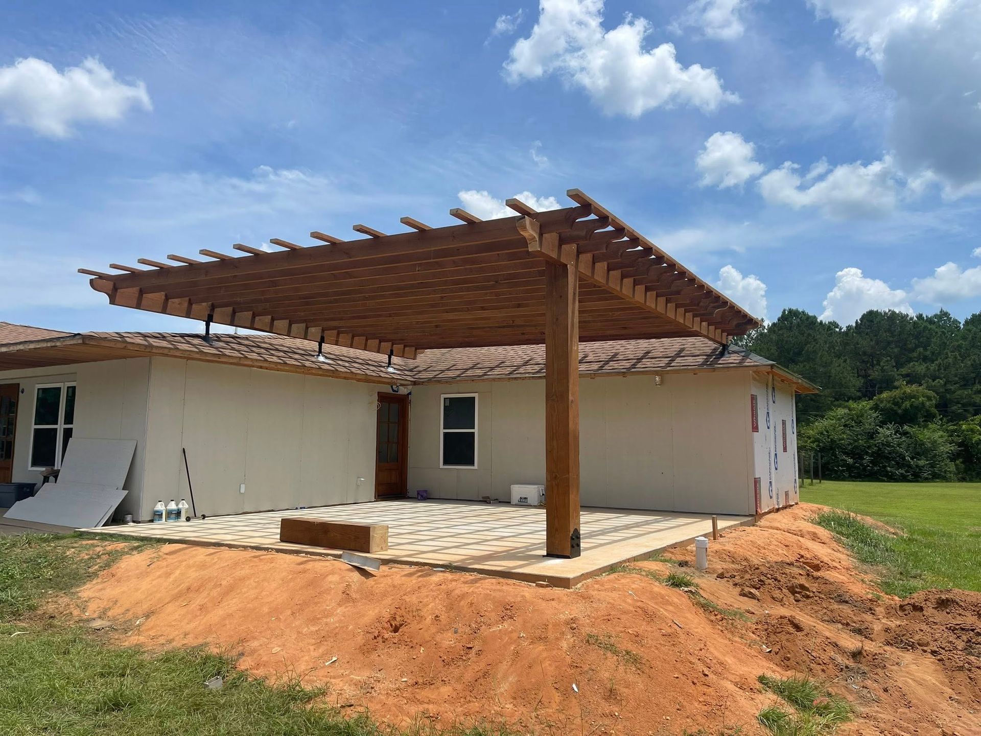 A house exterior under construction with a large wooden pergola covering a stone patio, set against a grassy landscape.