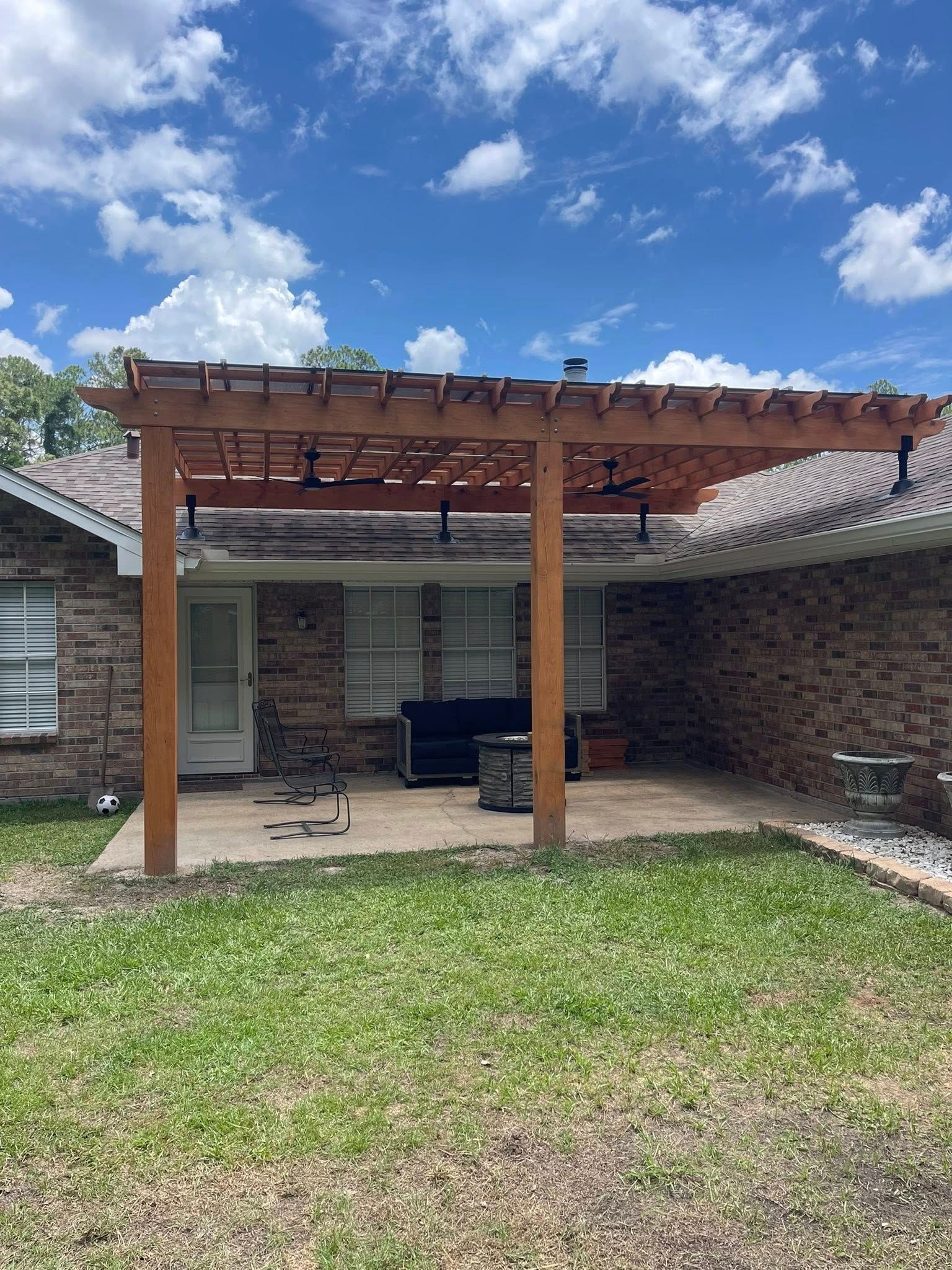 A wooden pergola attached to a brick house covers a patio with outdoor furniture against a blue, cloudy sky.