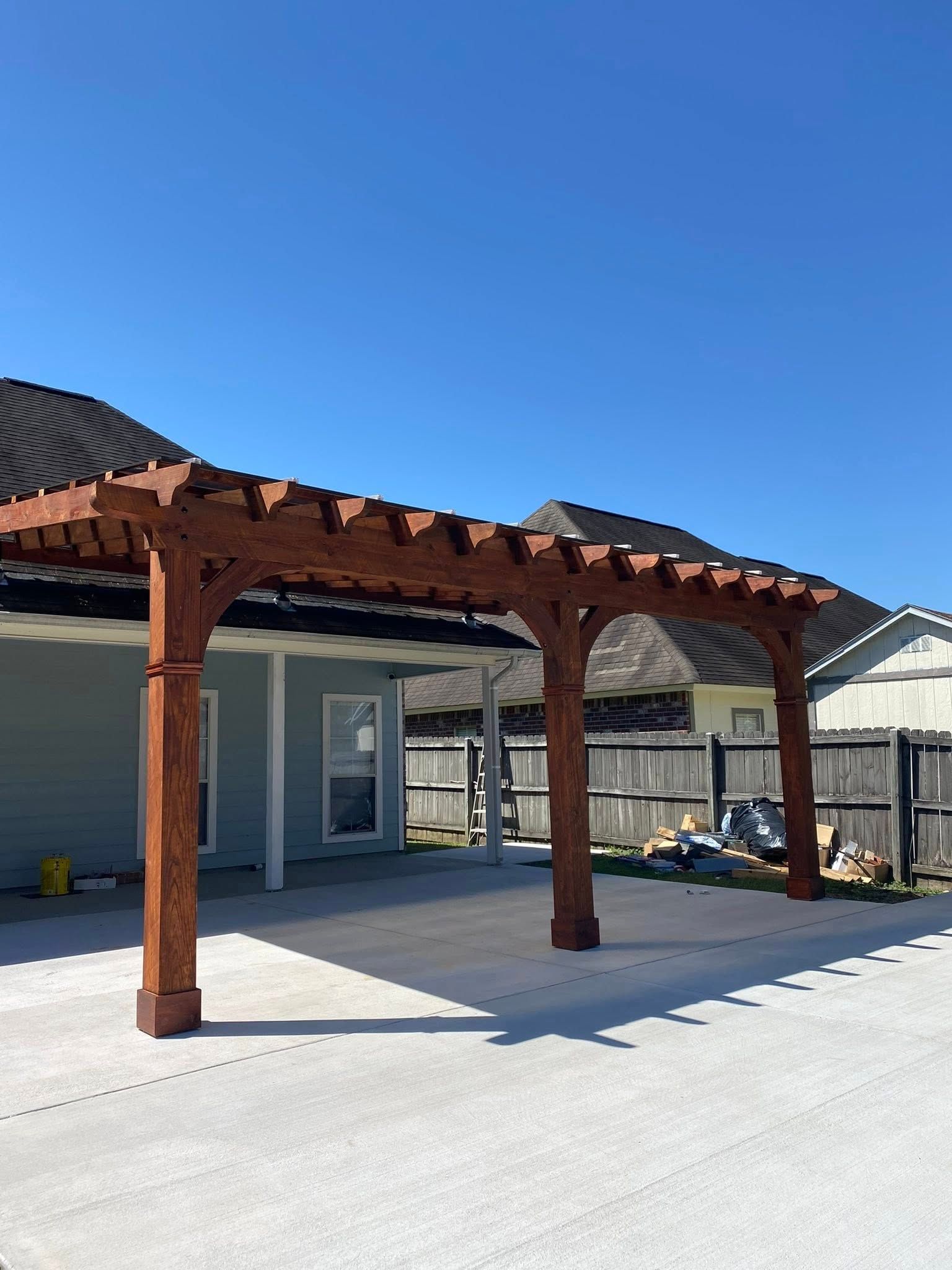 A wooden pergola stands on a concrete patio attached to a blue house against a clear blue sky.