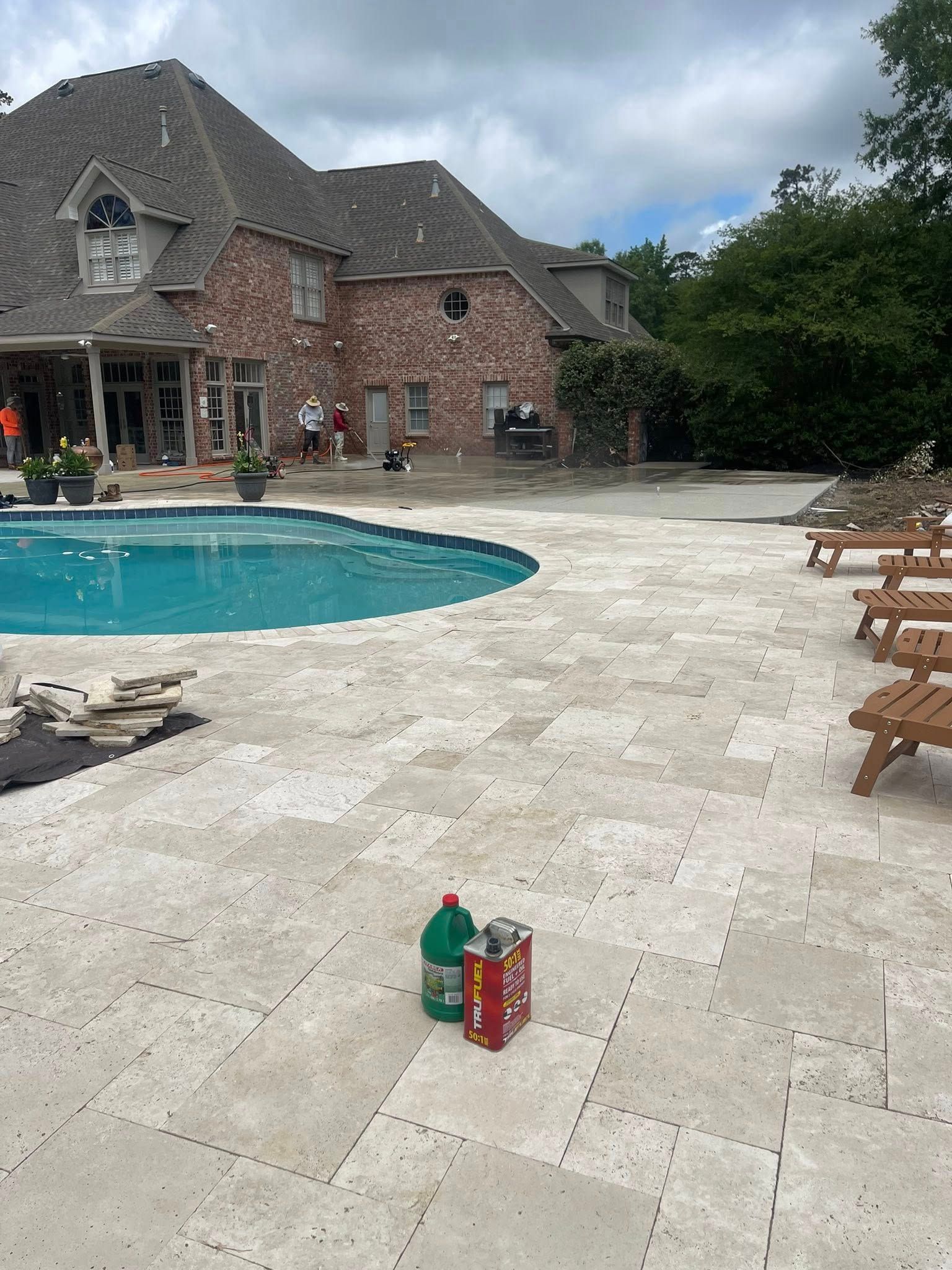 A brick house with a large stone pool deck and lounge chairs, featuring a green bottle and red can on the ground.