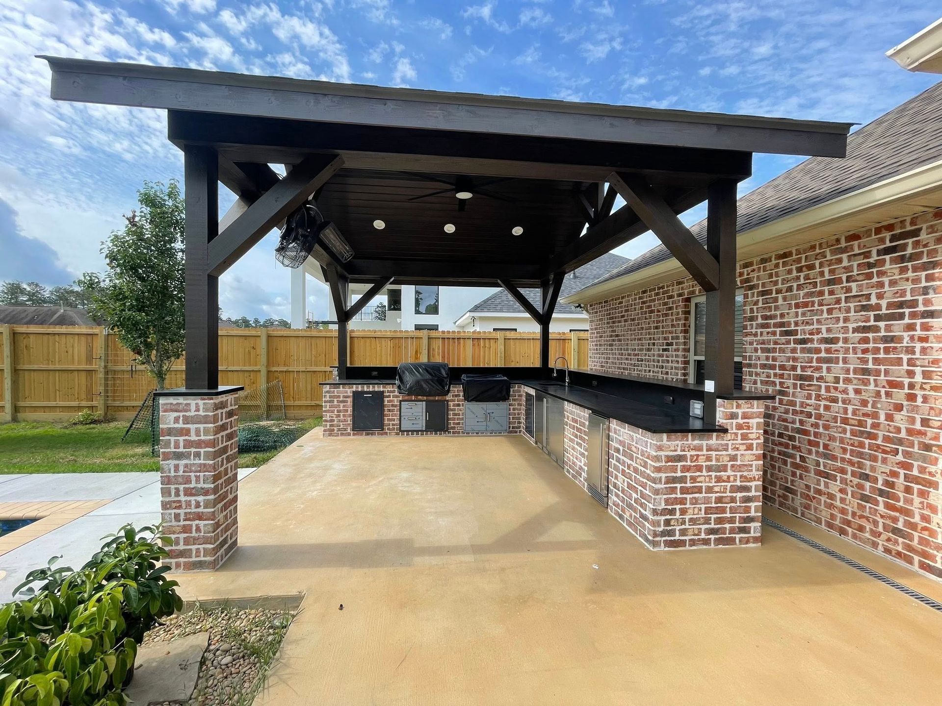 An outdoor kitchen under a wooden patio cover, featuring brick columns and a black counter, attached to a brick house.