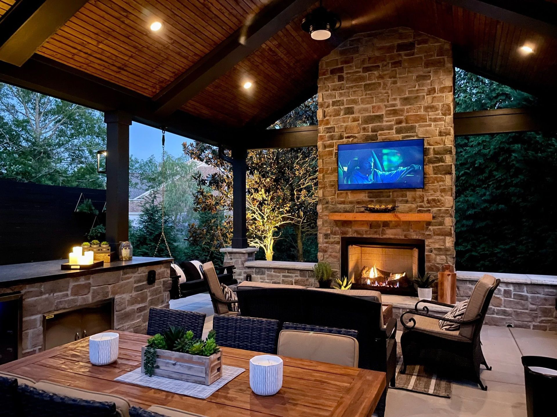 An outdoor covered patio with a stone fireplace, television, dining table, and seating area at dusk.