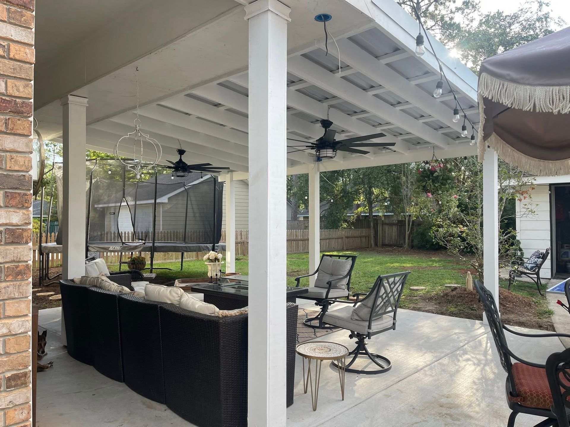 A covered backyard patio with outdoor furniture, including a couch, chairs, and two ceiling fans, overlooking a lawn.