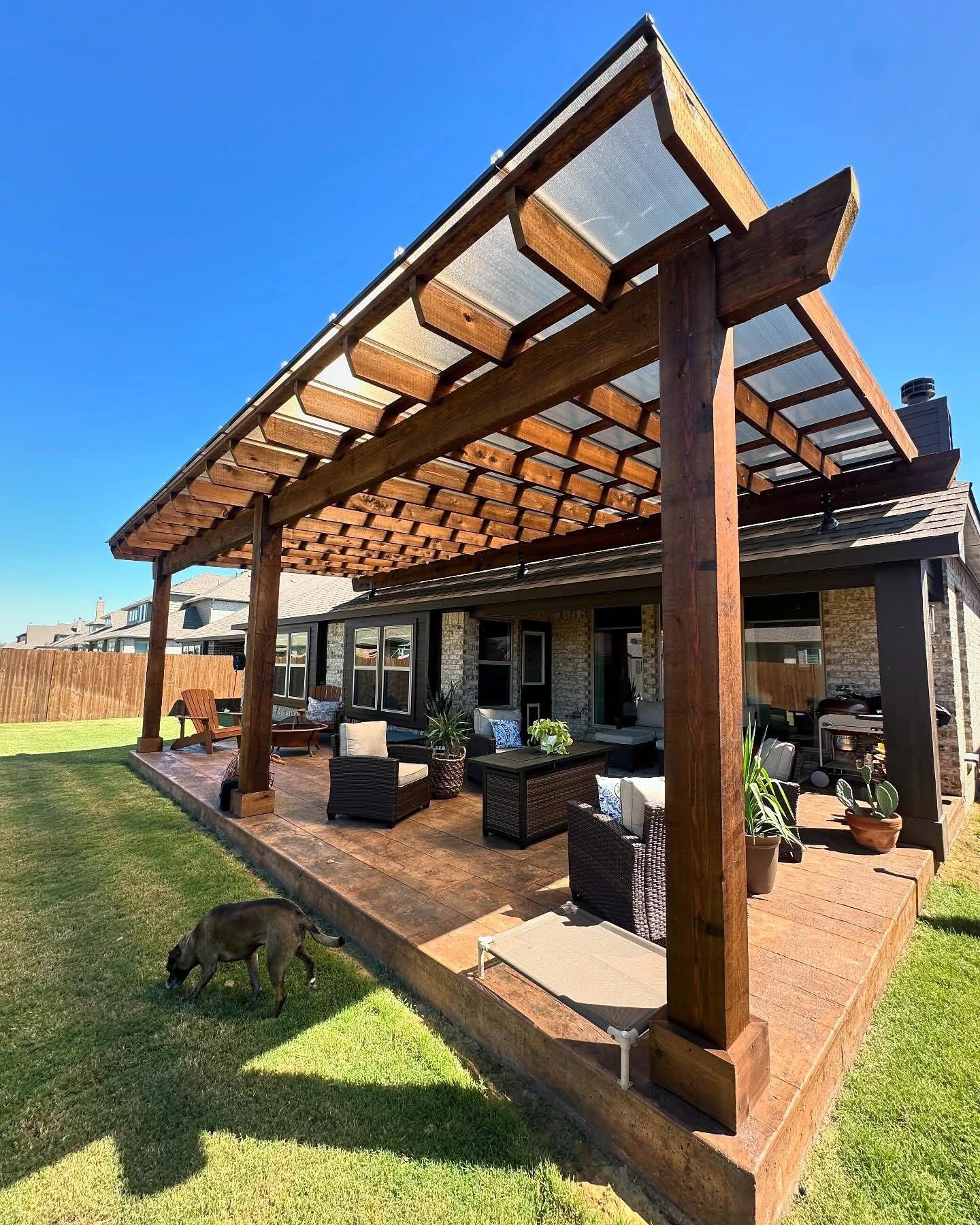 A wooden pergola with a translucent roof shades a furnished patio, while a dog walks on the grass in the foreground.