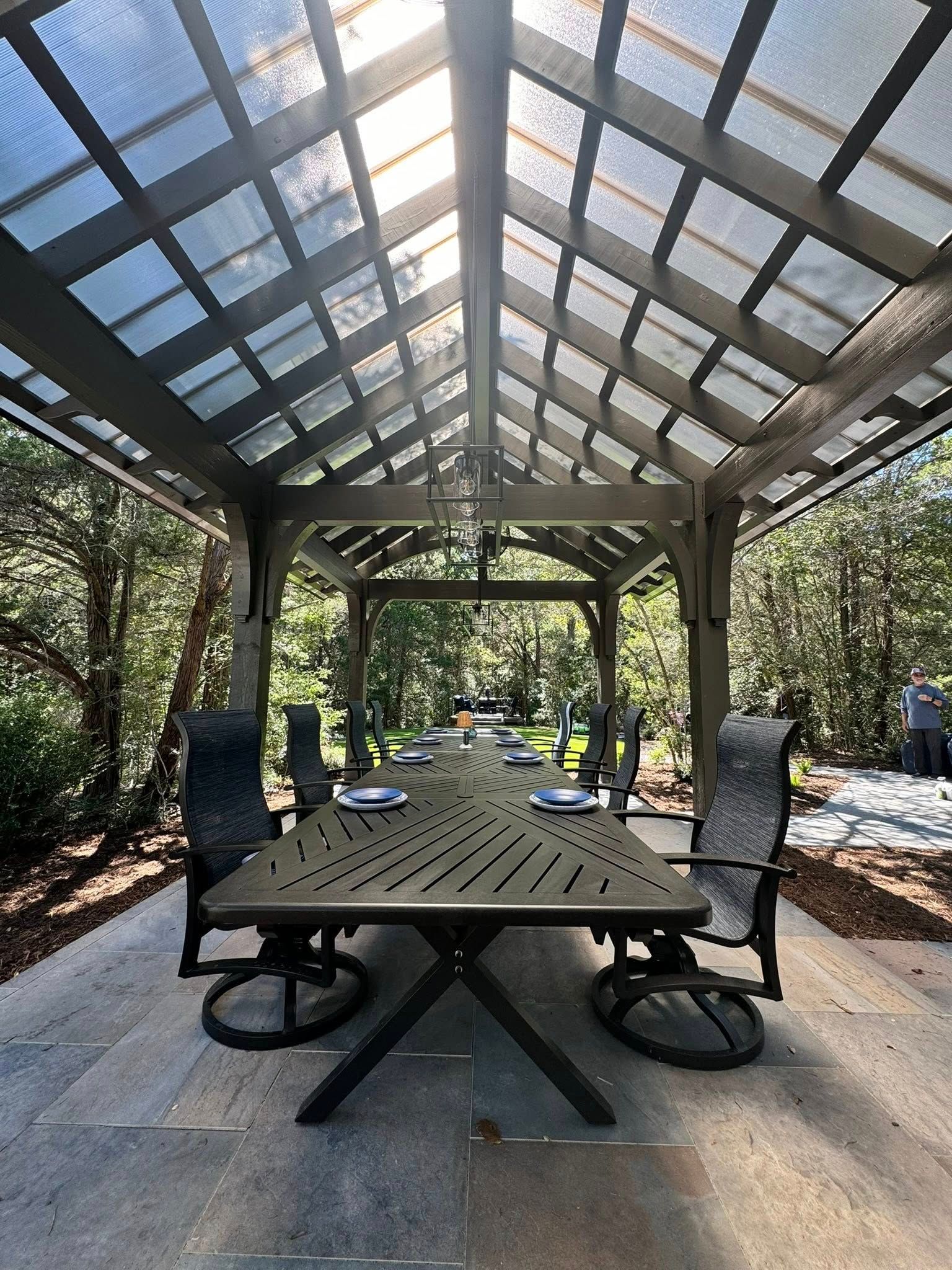 An outdoor dining table with chairs under a wooden pergola roof on a stone patio, surrounded by trees and greenery.
