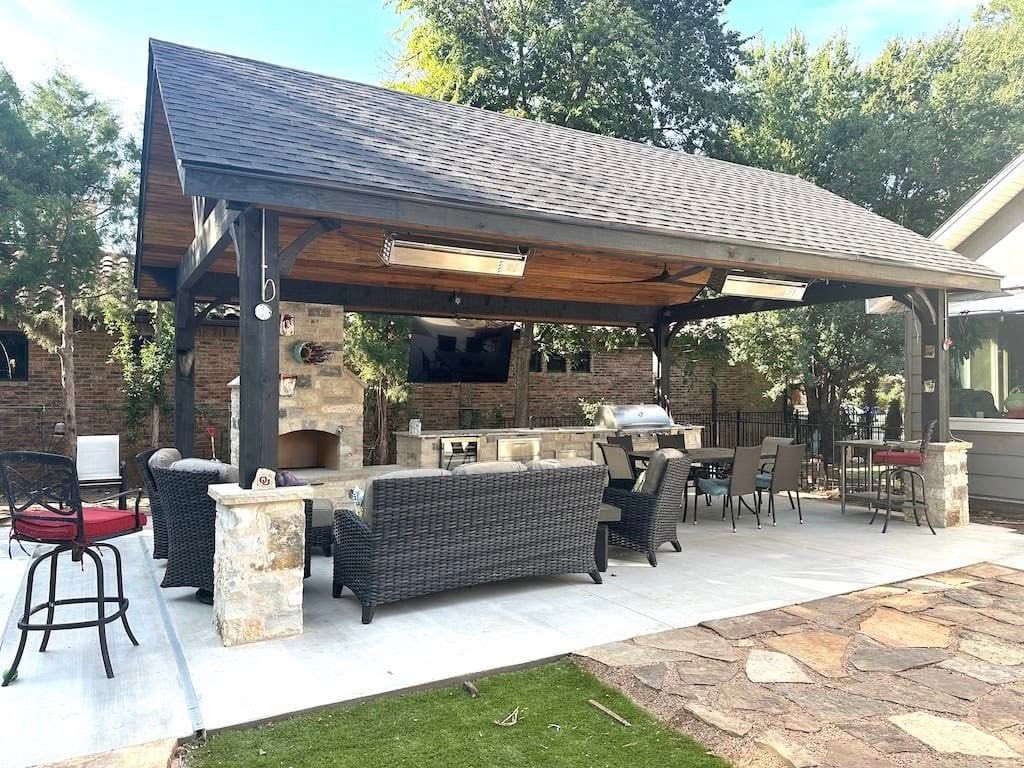An outdoor patio with a large covered pavilion, stone fireplace, cushioned furniture, and a grill set on a concrete floor.