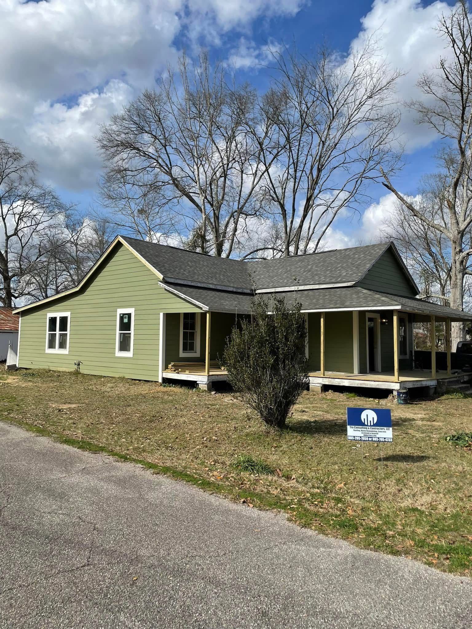 A light green, single-story house with a grey roof and wraparound porch sits on a grassy lot under a blue, cloudy sky.