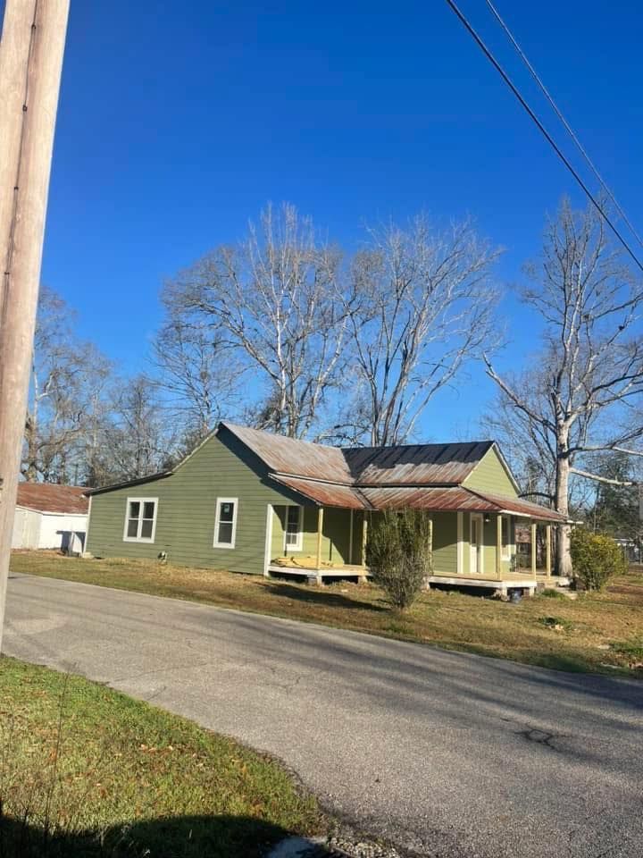 A sage green, single-story house with a rusted metal roof and a front porch sits along a street under a clear blue sky.
