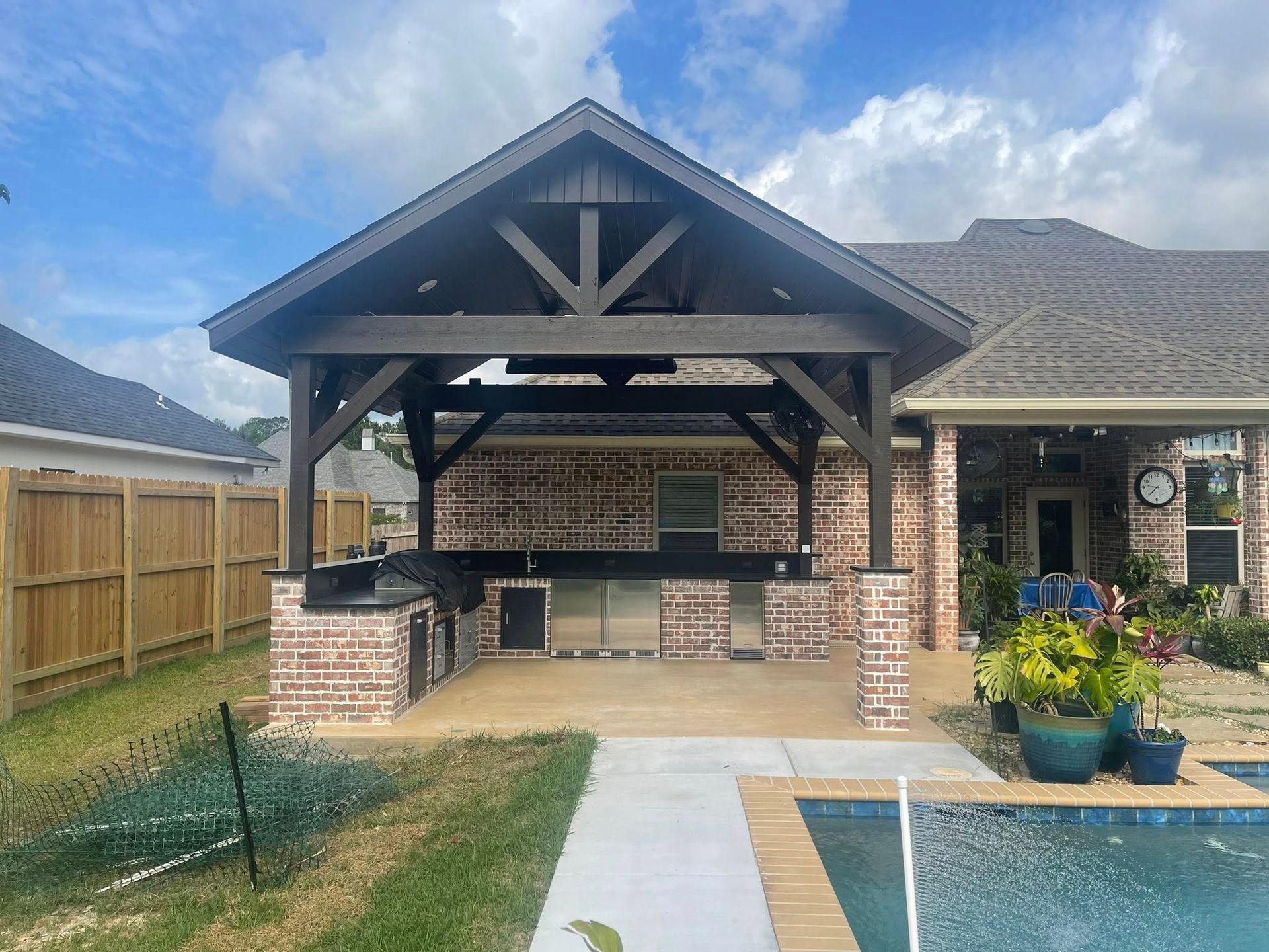 A covered outdoor kitchen with brick counters and dark wood beams, situated in a backyard beside a swimming pool.