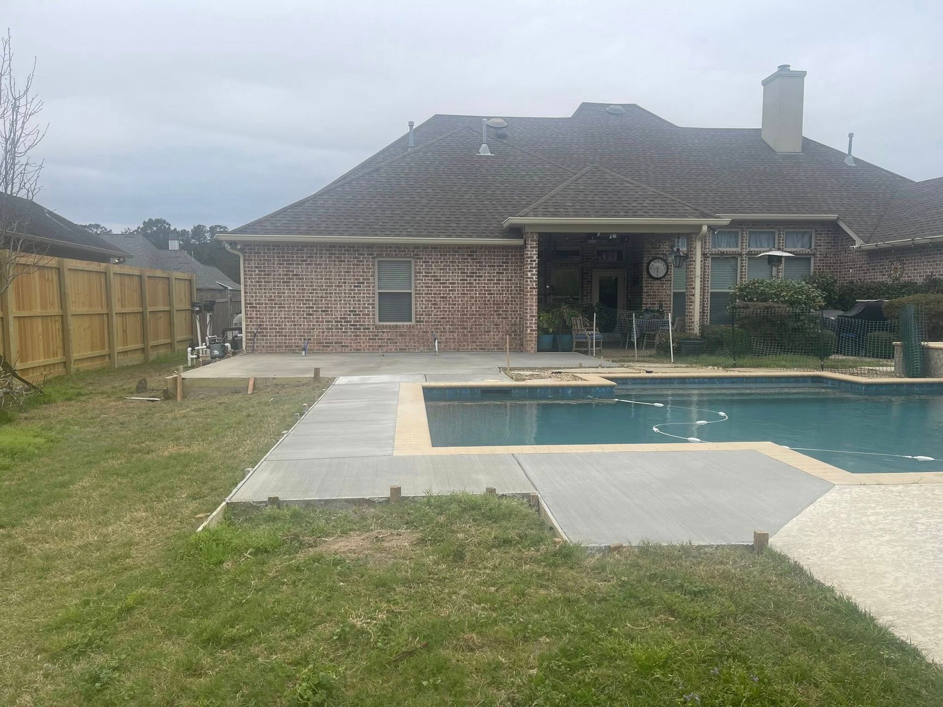 A brick house with a newly poured concrete pool deck and patio area in a backyard with a wooden fence.