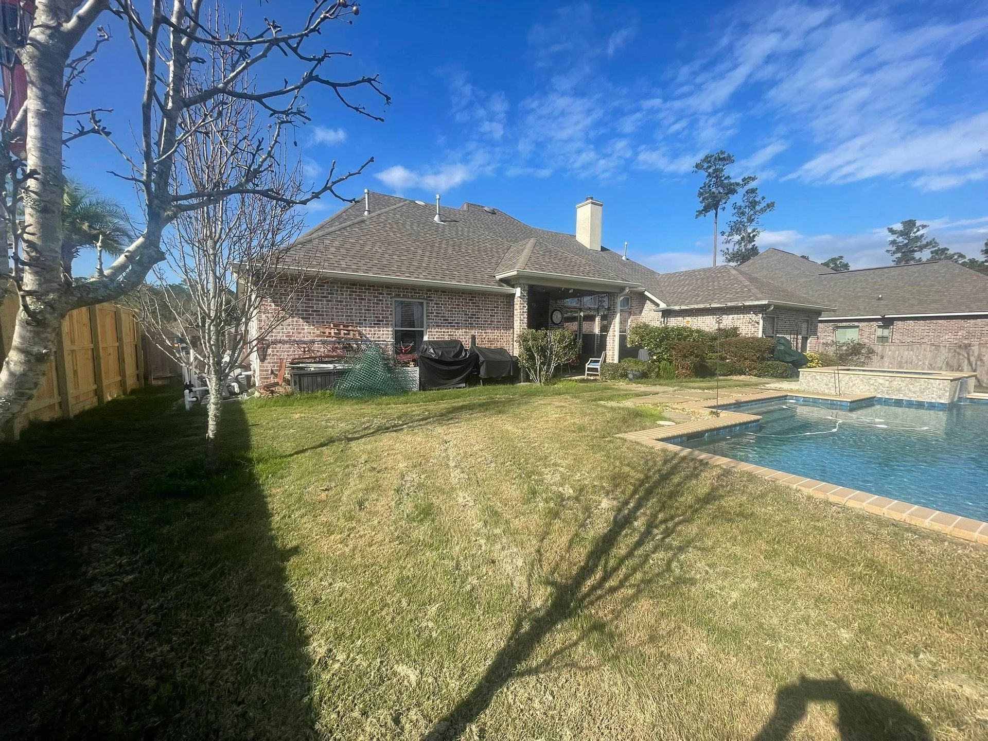 A sunny backyard featuring a brick house, a swimming pool, and a lawn with tree shadows cast across the grass.