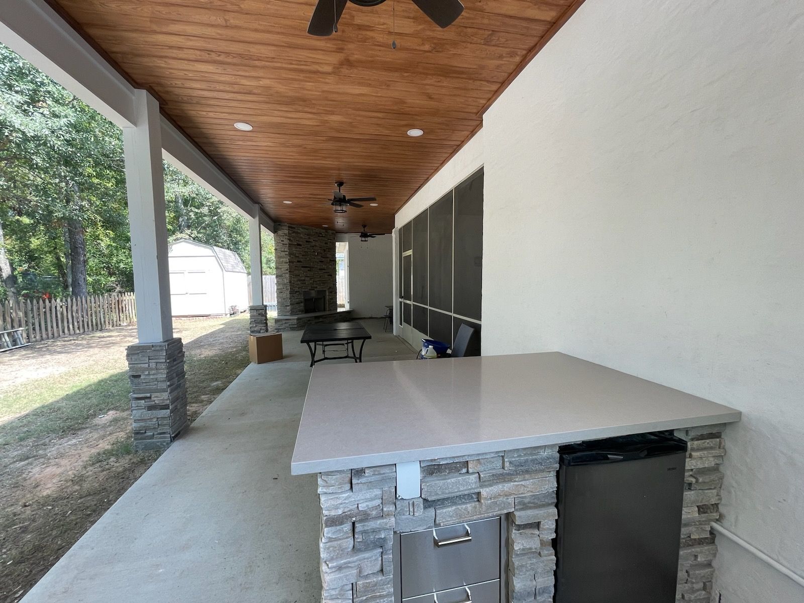 A covered outdoor patio with a stone-base bar counter, a stainless steel drawer, a small refrigerator, and wood ceiling.