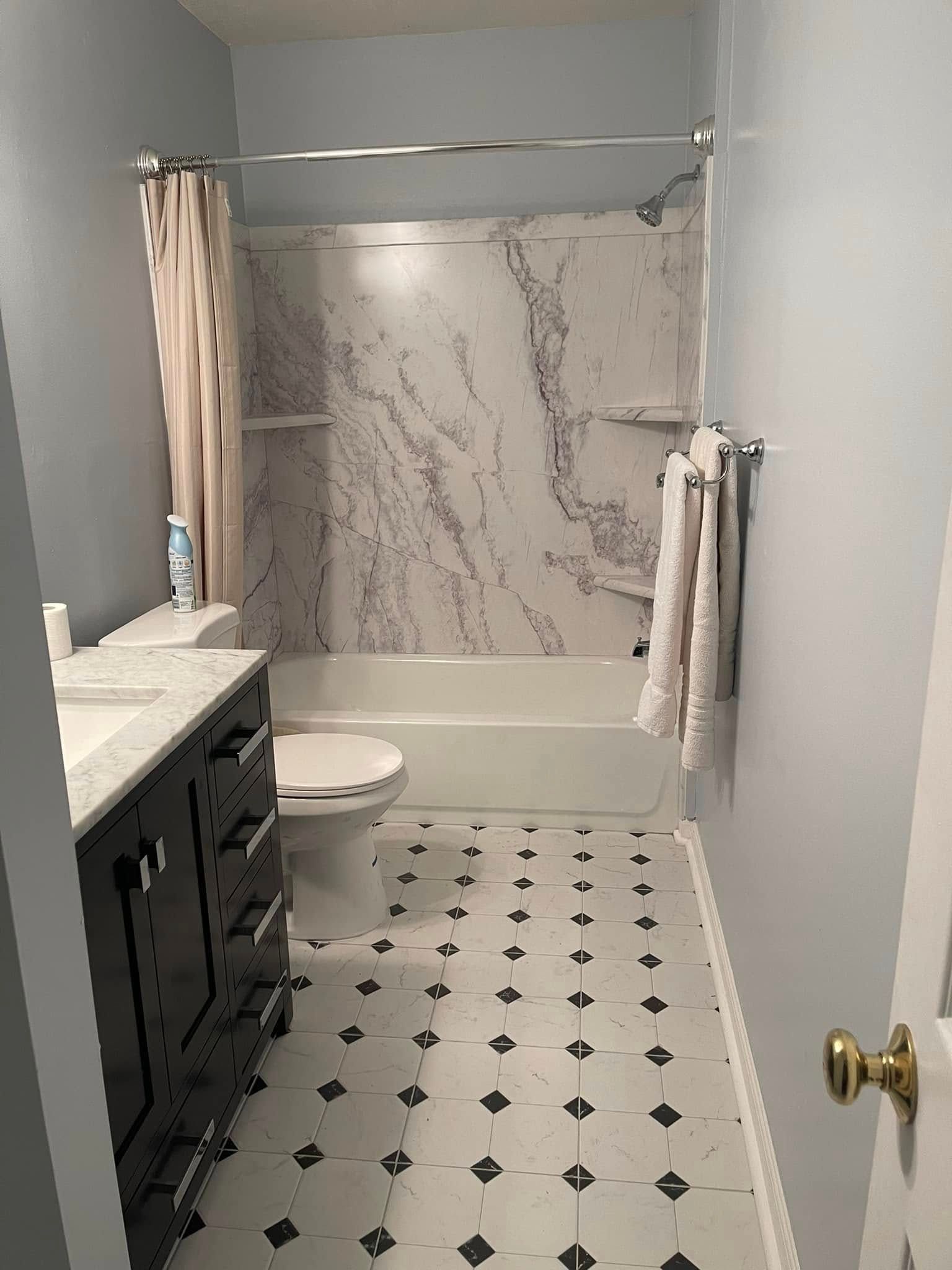 A bathroom with white walls, a black vanity, marble-patterned shower surround, and white tiled floor with black accents.