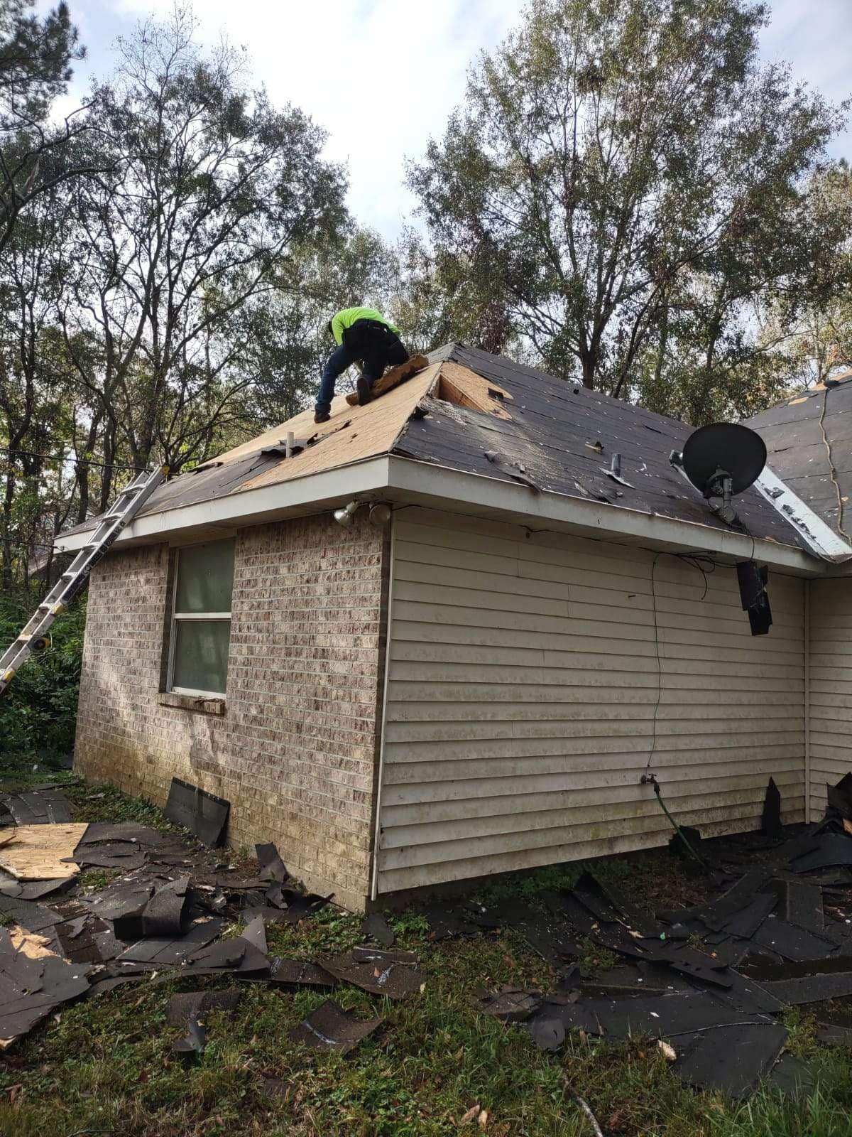 A person in a high-visibility vest repairs the roof of a beige house with debris scattered on the ground below.