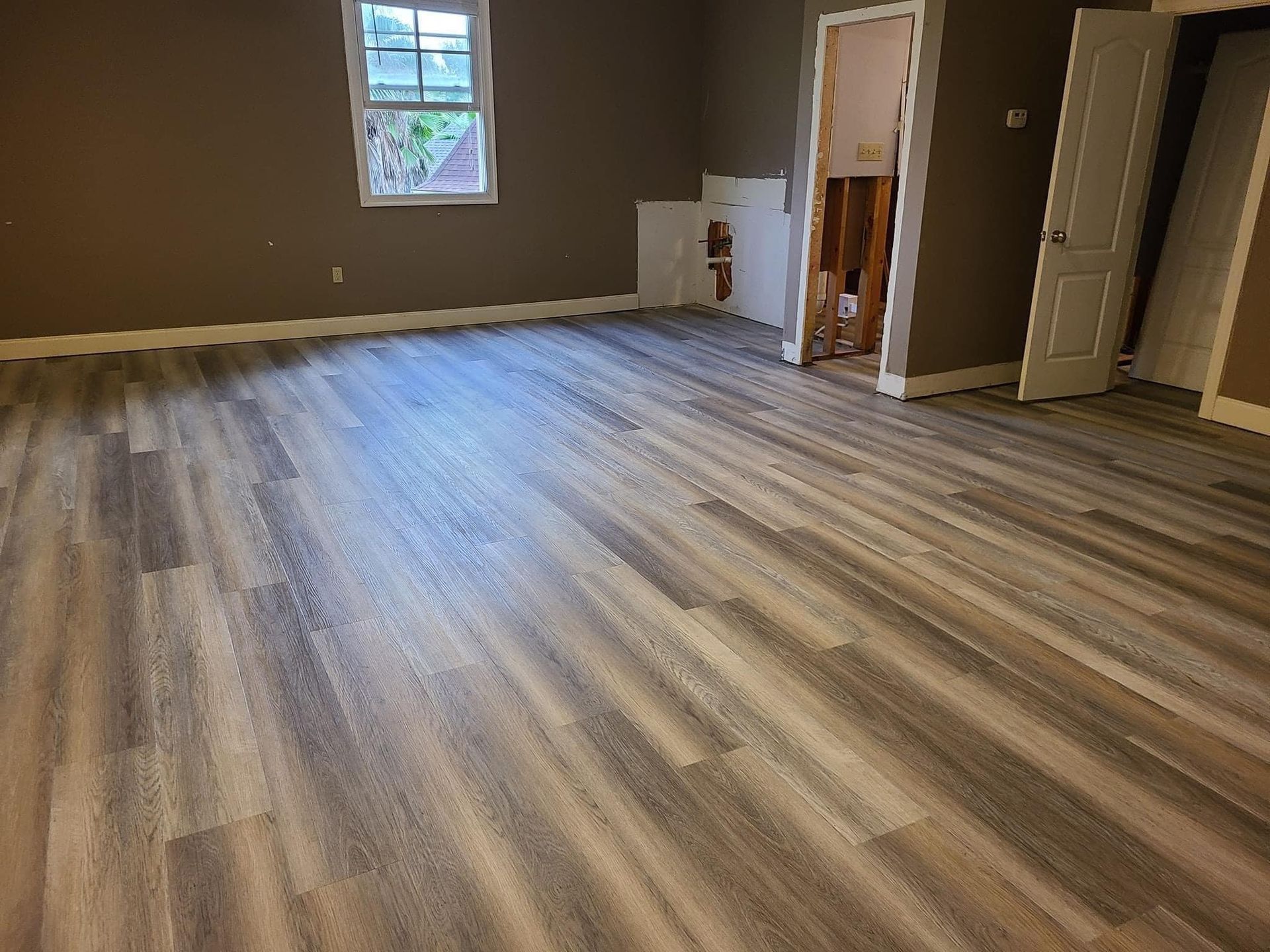 A room with wood-look laminate flooring, dark grey walls, a single window, and an open doorway leading to another room.