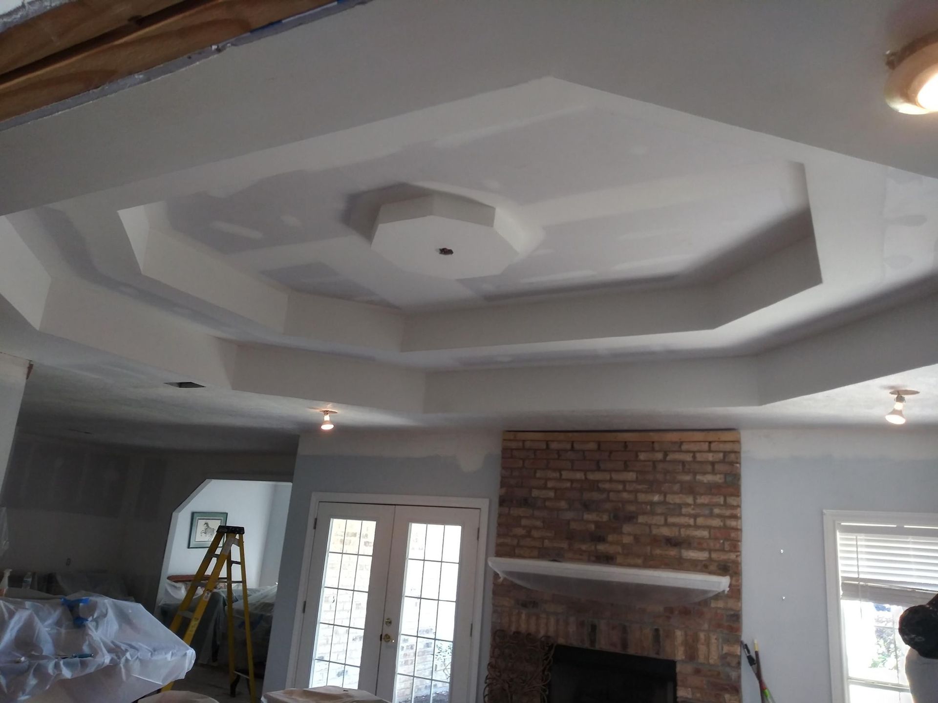 A room featuring a white multi-tiered octagonal tray ceiling, a brick fireplace, and a yellow step ladder.