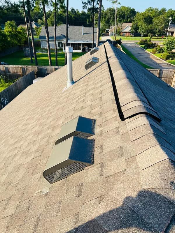 A view of a brown shingled residential roof with metal vents, a chimney pipe, and a ridge vent under a clear blue sky.