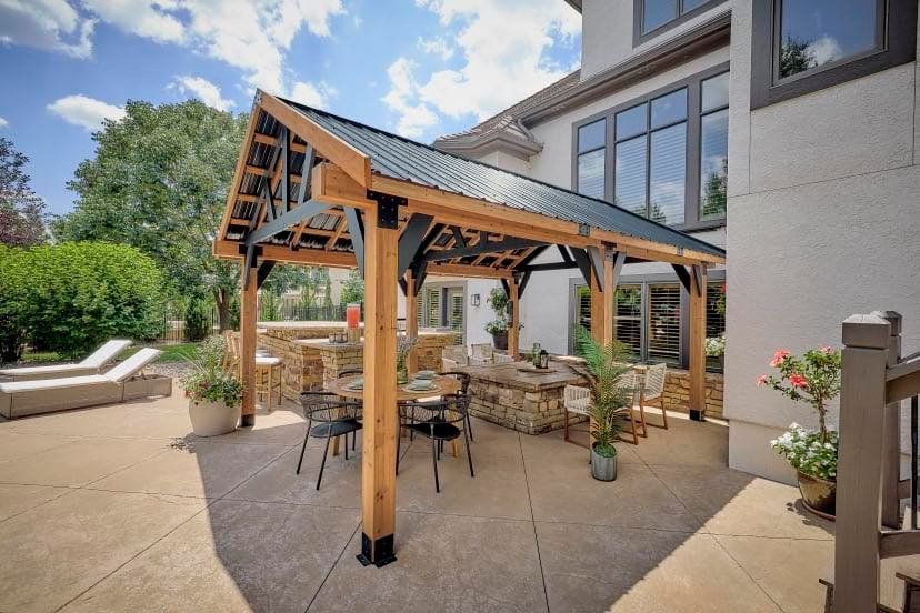 A wooden pavilion with a dark metal roof stands over an outdoor dining area on a patio next to a house.