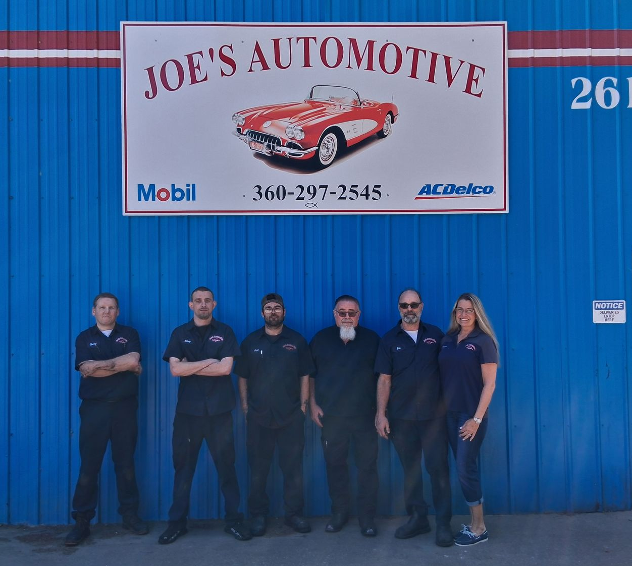 Six people standing outside Joe’s Automotive under a sign on a blue building.