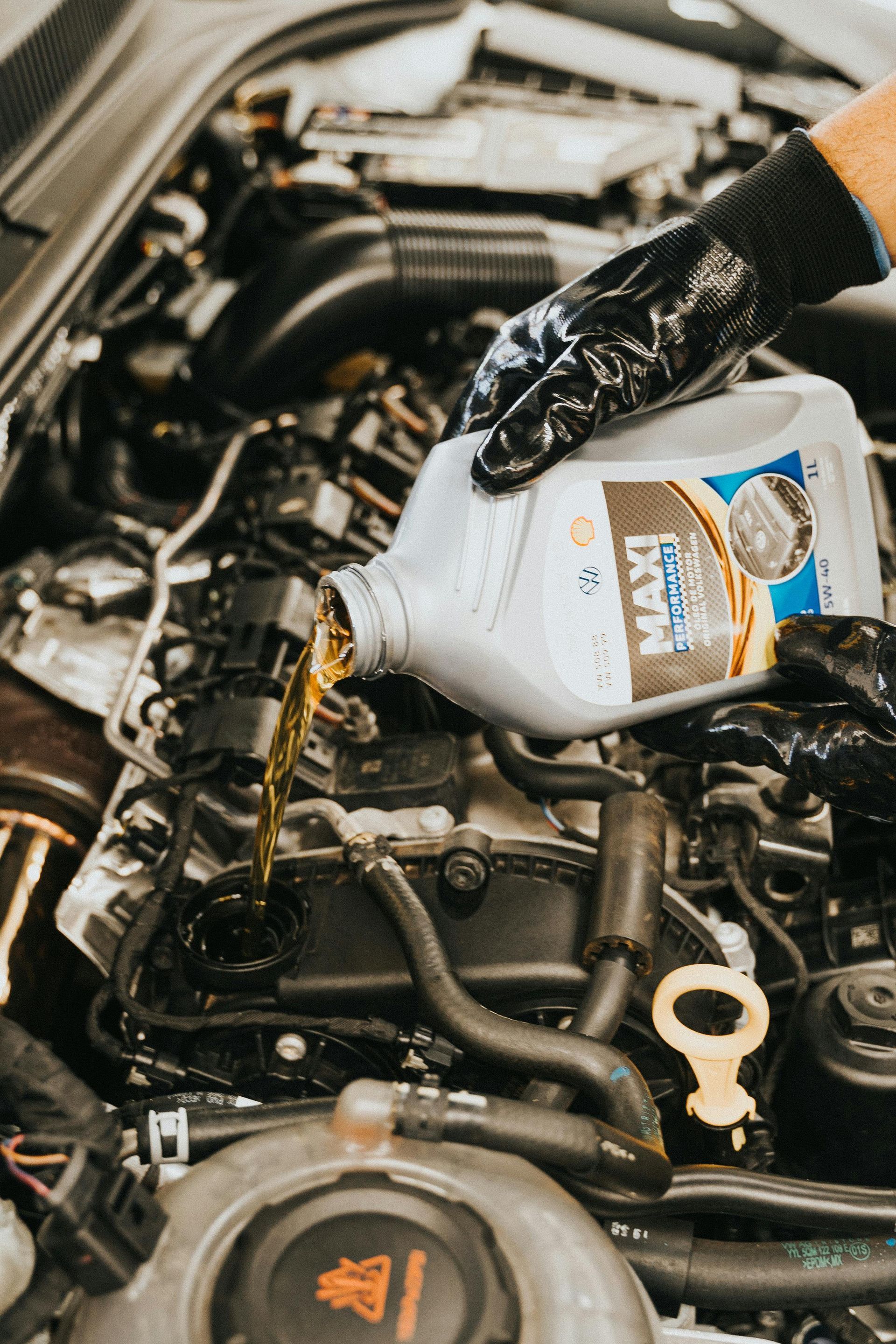 Gloved hand pouring oil from a white bottle into a car engine compartment