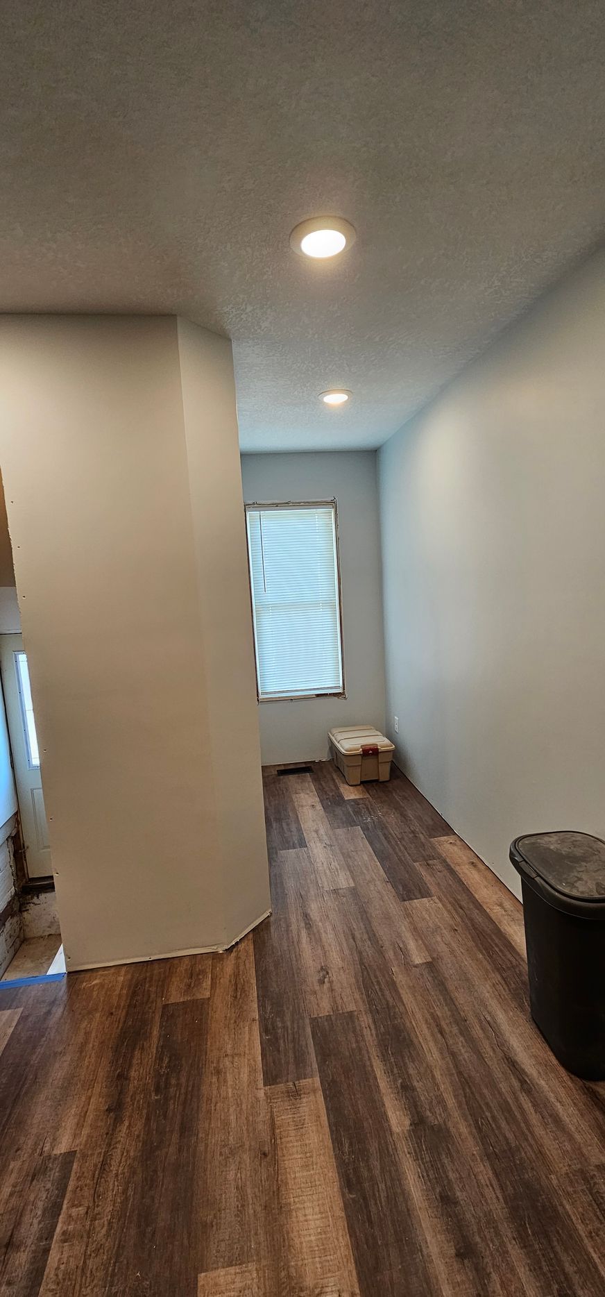 Empty hallway with wood floors, white walls, recessed ceiling lights, and a window at the end.