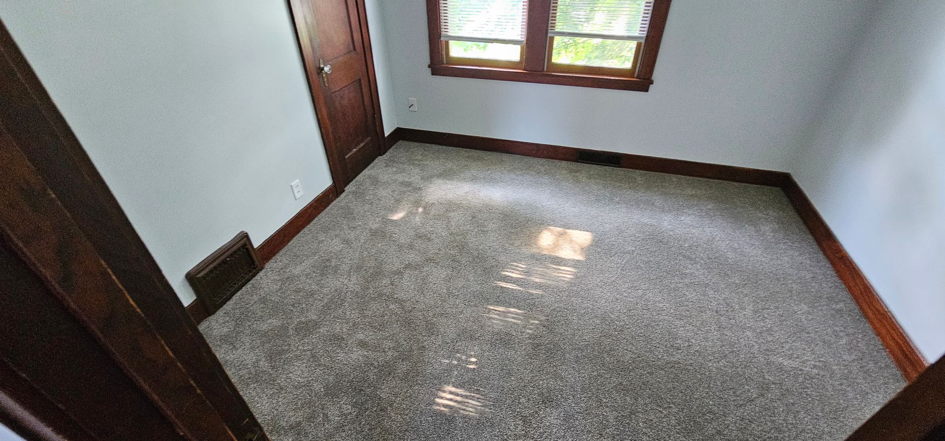 Empty carpeted room with white walls, dark wood trim, and a window letting in light.