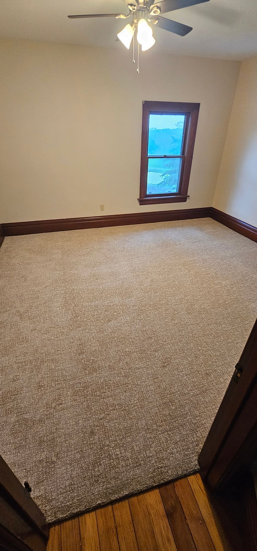 Empty carpeted bedroom with beige walls, a ceiling fan, and one small window.