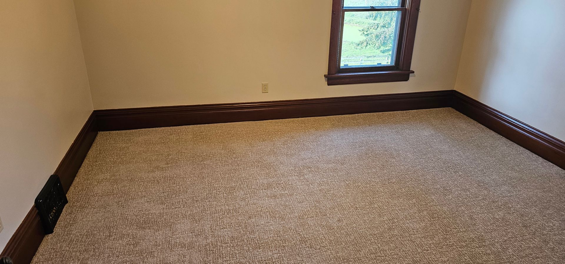 Empty beige carpeted room with tan walls, dark trim, and a small window.