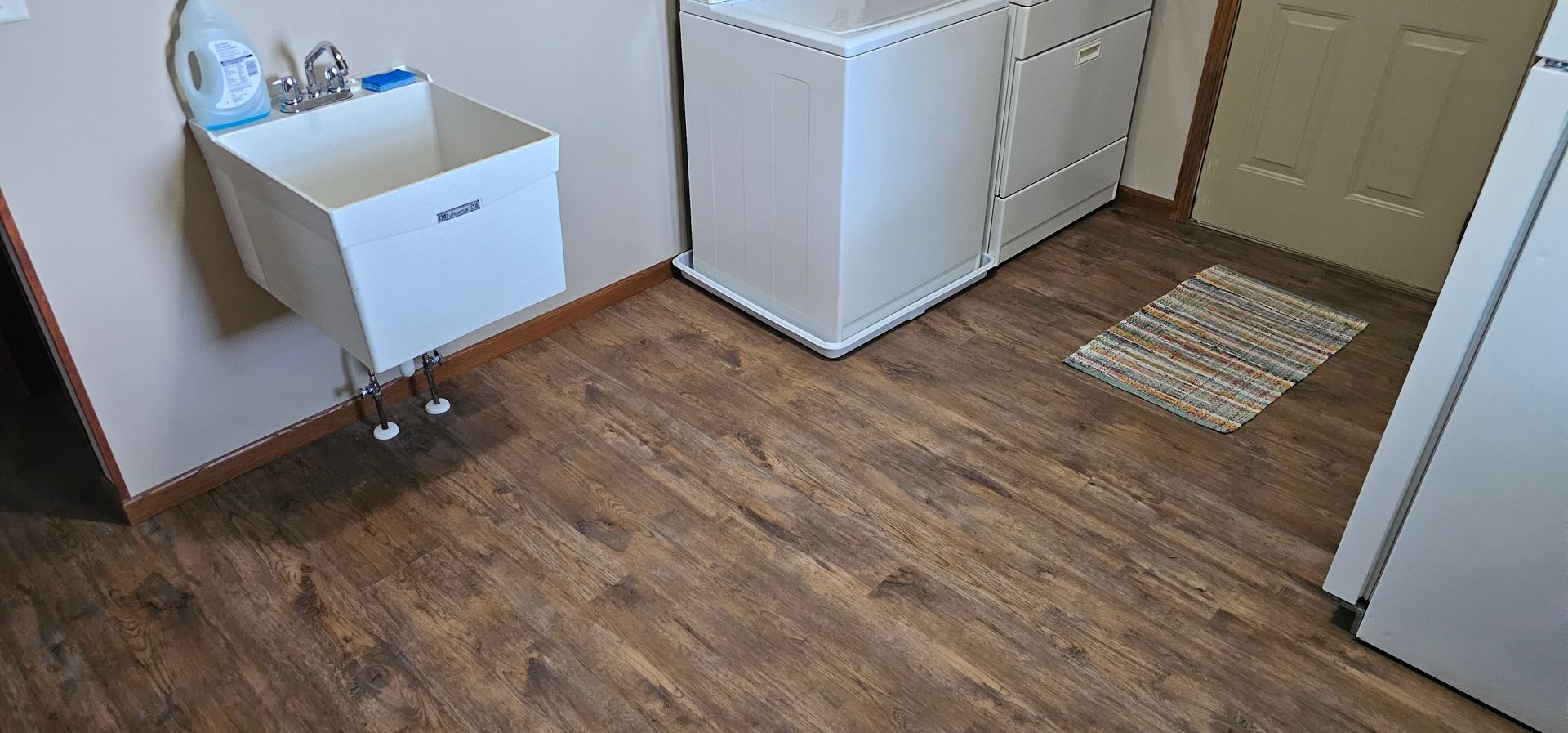 Small laundry room with wood-look flooring, white cabinet, sink, and a beige door with a rug.