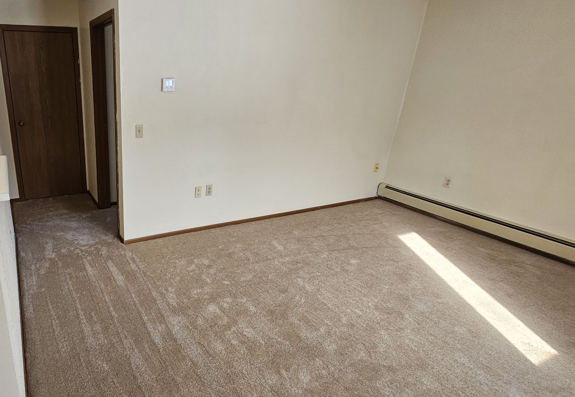 Empty beige room with carpet, open doorway, and sunlight streaming across the floor.