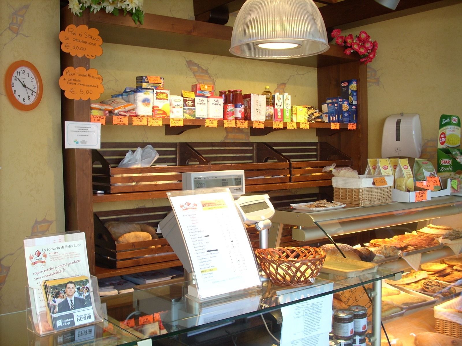 Bancone panetteria con pasticcini, scaffali per il pane e cartelli con il menù, il tutto illuminato da una luce calda.