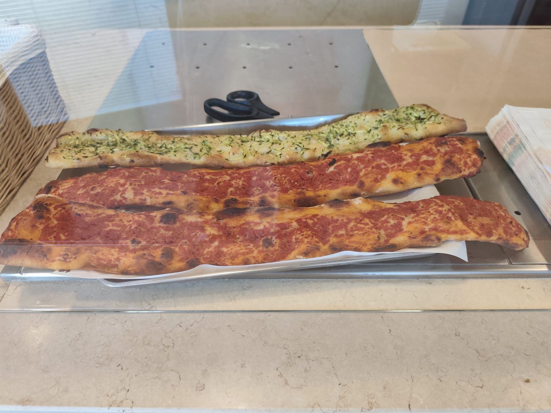 Tre focacce rettangolari lunghe, una con la superficie verde e due con la superficie rossa, su un bancone dietro una vetrina.