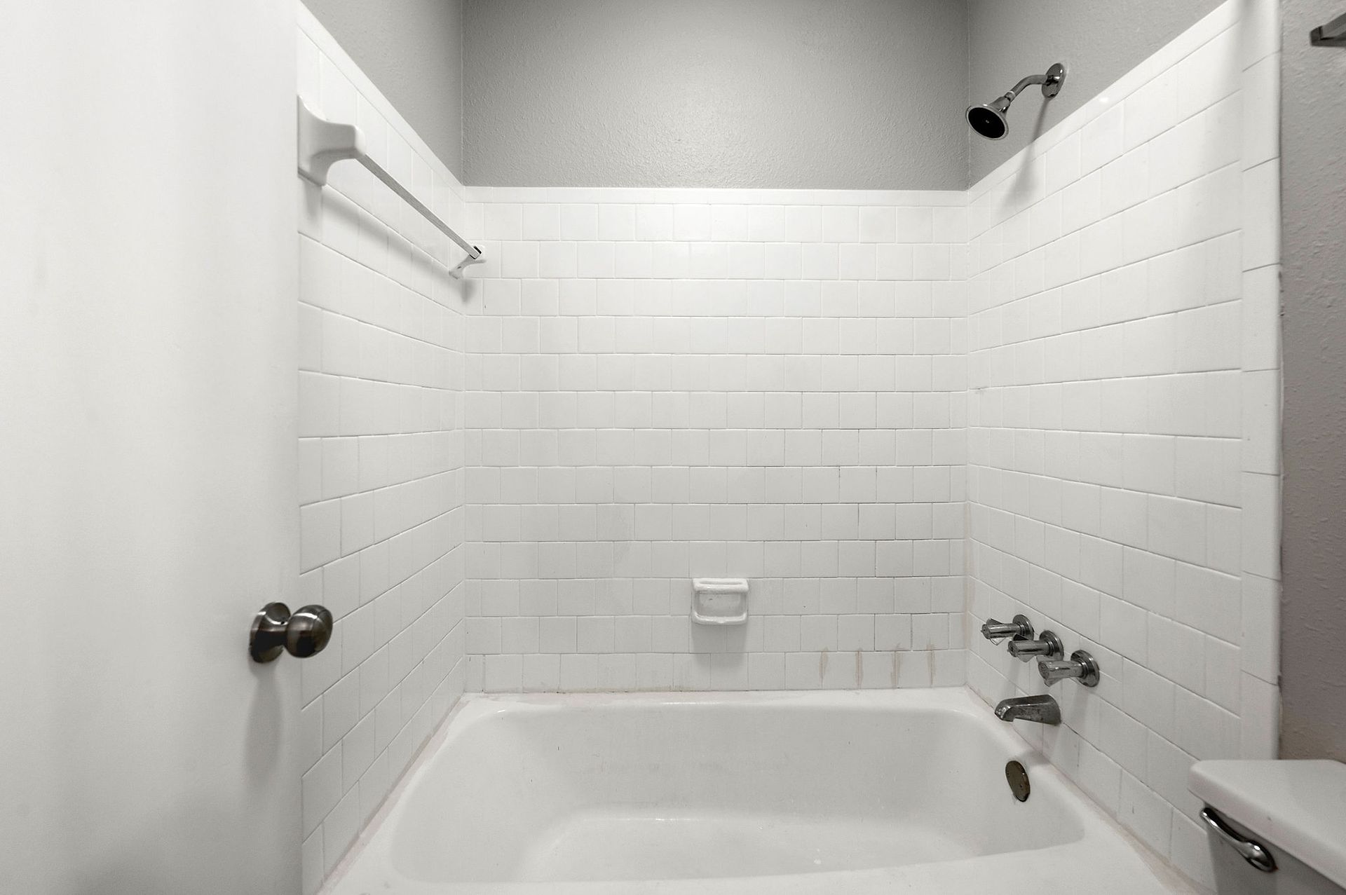 White-tiled bathtub/shower with silver fixtures and a small window above.