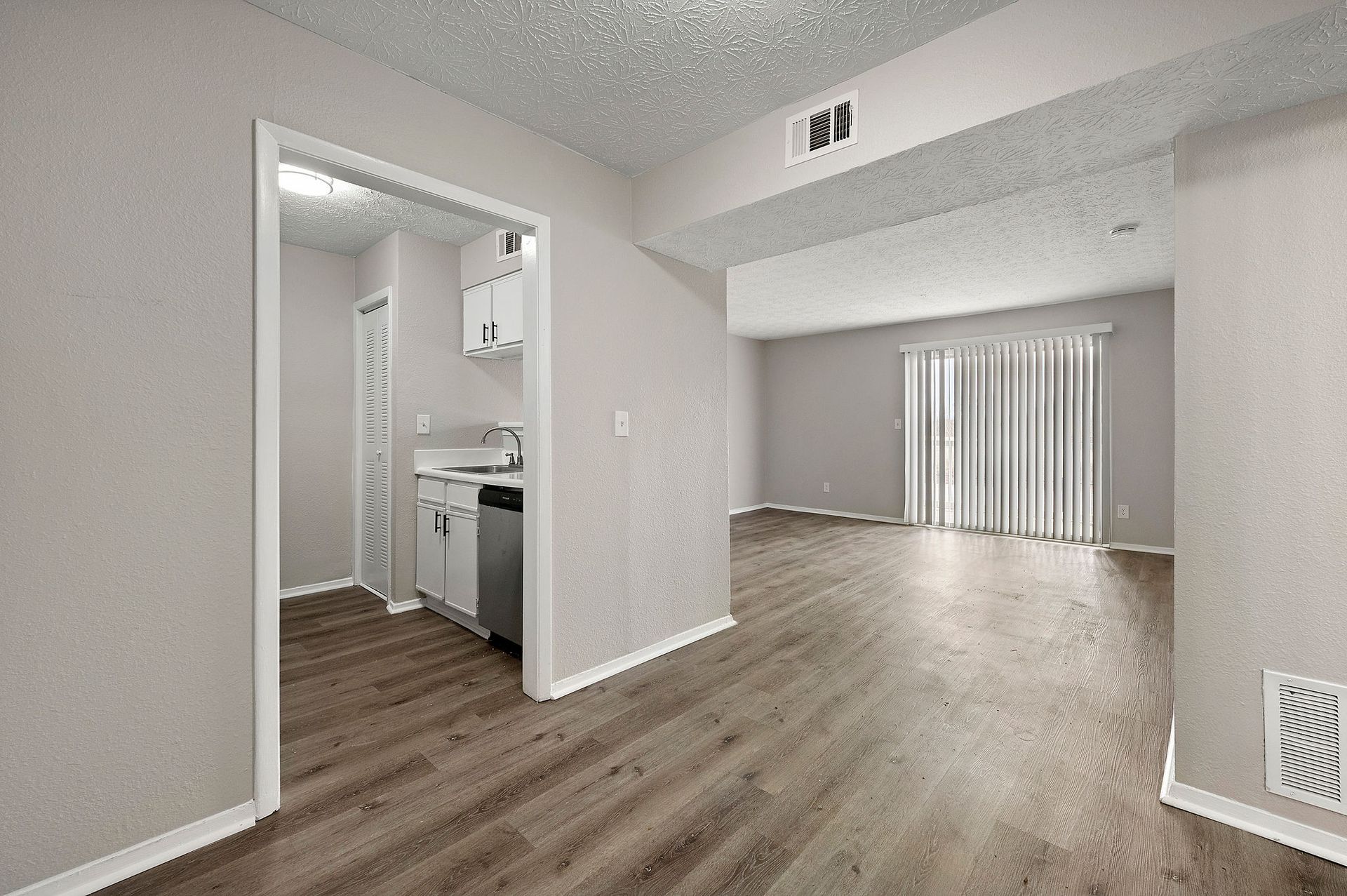 Empty apartment living room with wood floors, open kitchen, and sliding glass doors with vertical blinds