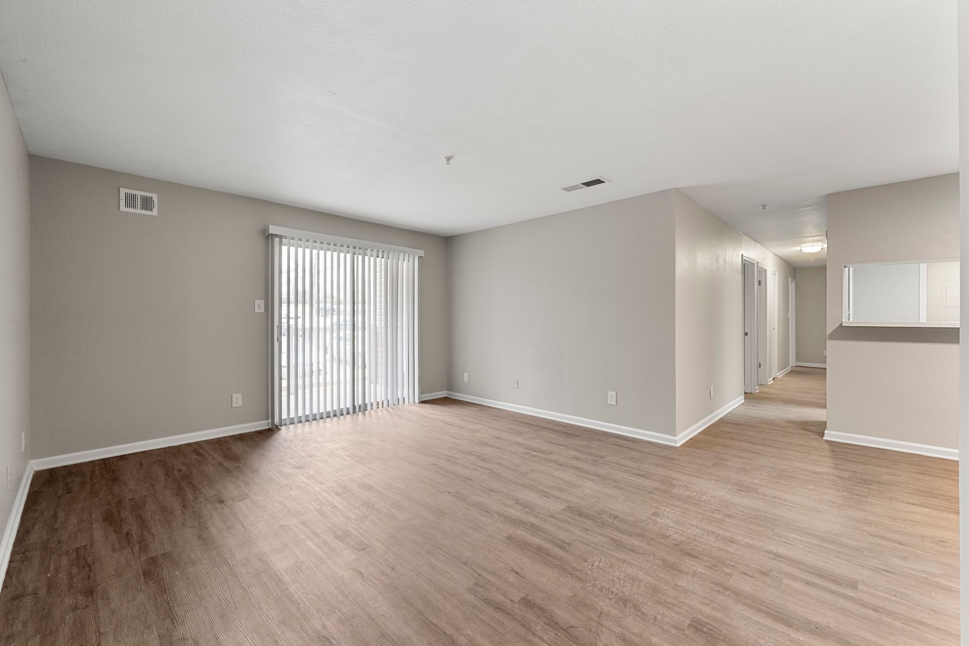 Empty living room with light wood floors, gray walls, sliding glass door, and hallway to kitchen