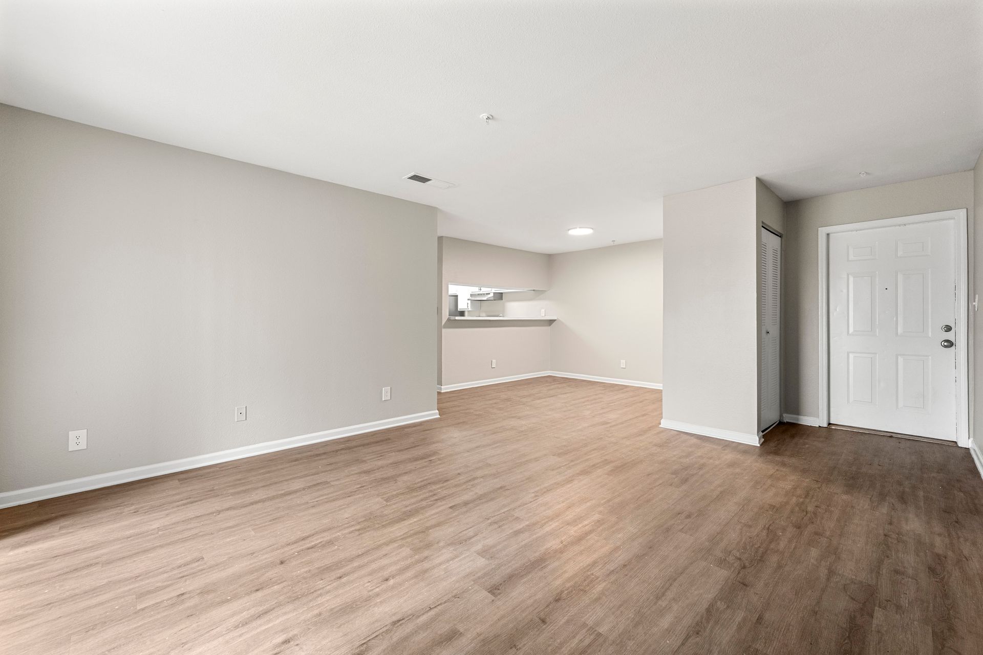 Empty living room with light wood floors, beige walls, and a view into a kitchen area.