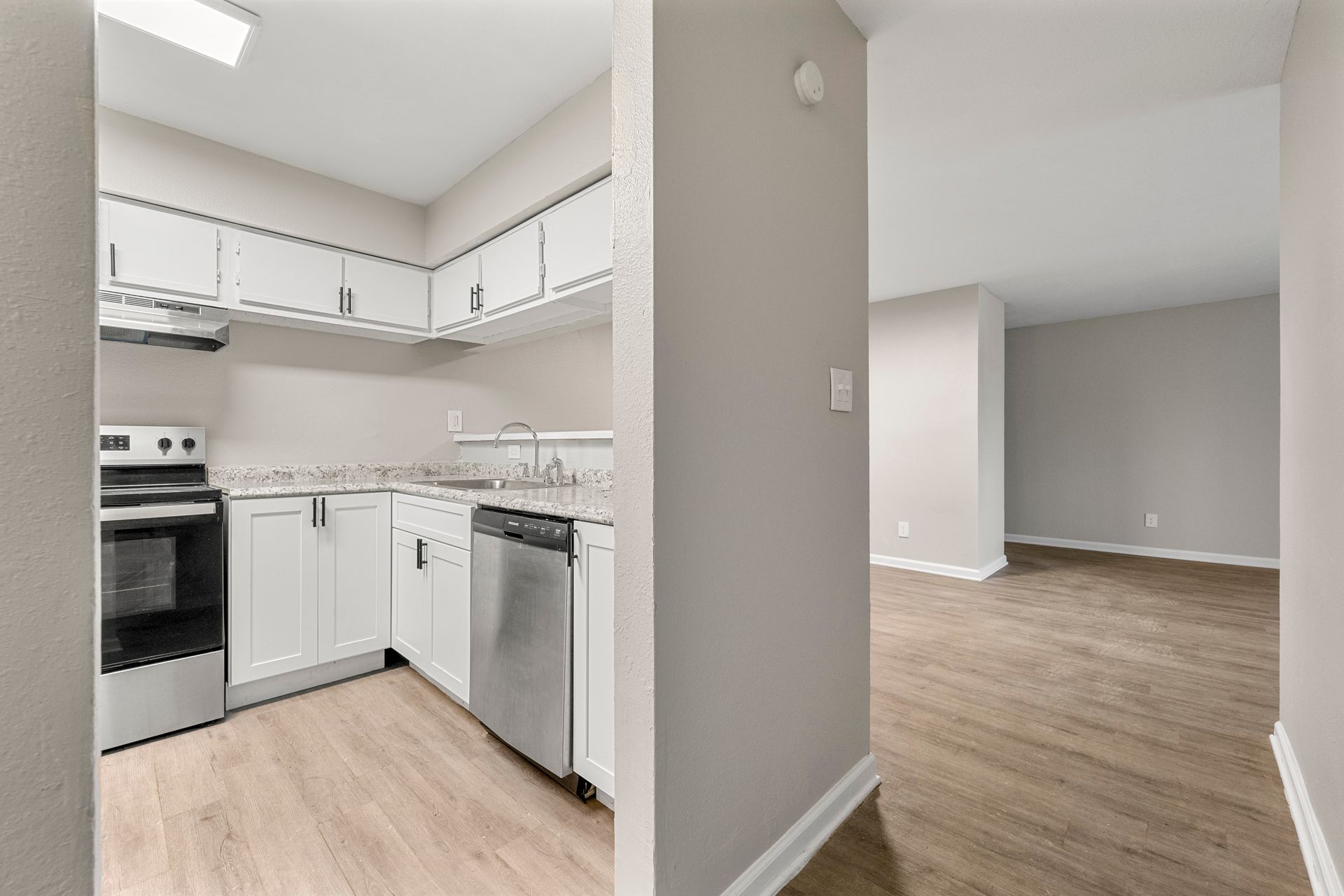 Bright kitchen with white cabinets and stainless appliances opening to an empty gray living room.