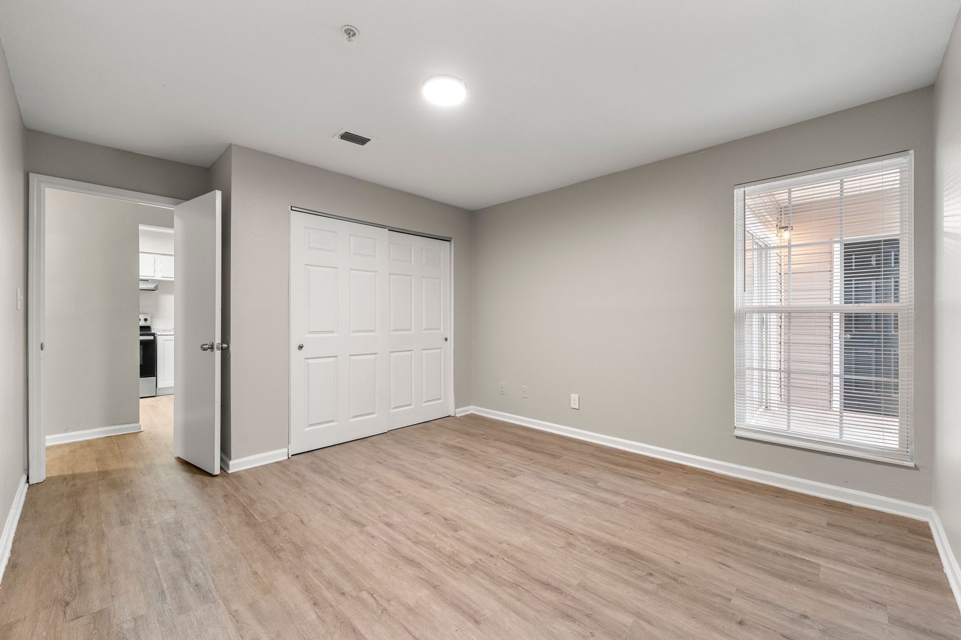 Empty beige bedroom with wood flooring, closet doors, and a window with white blinds