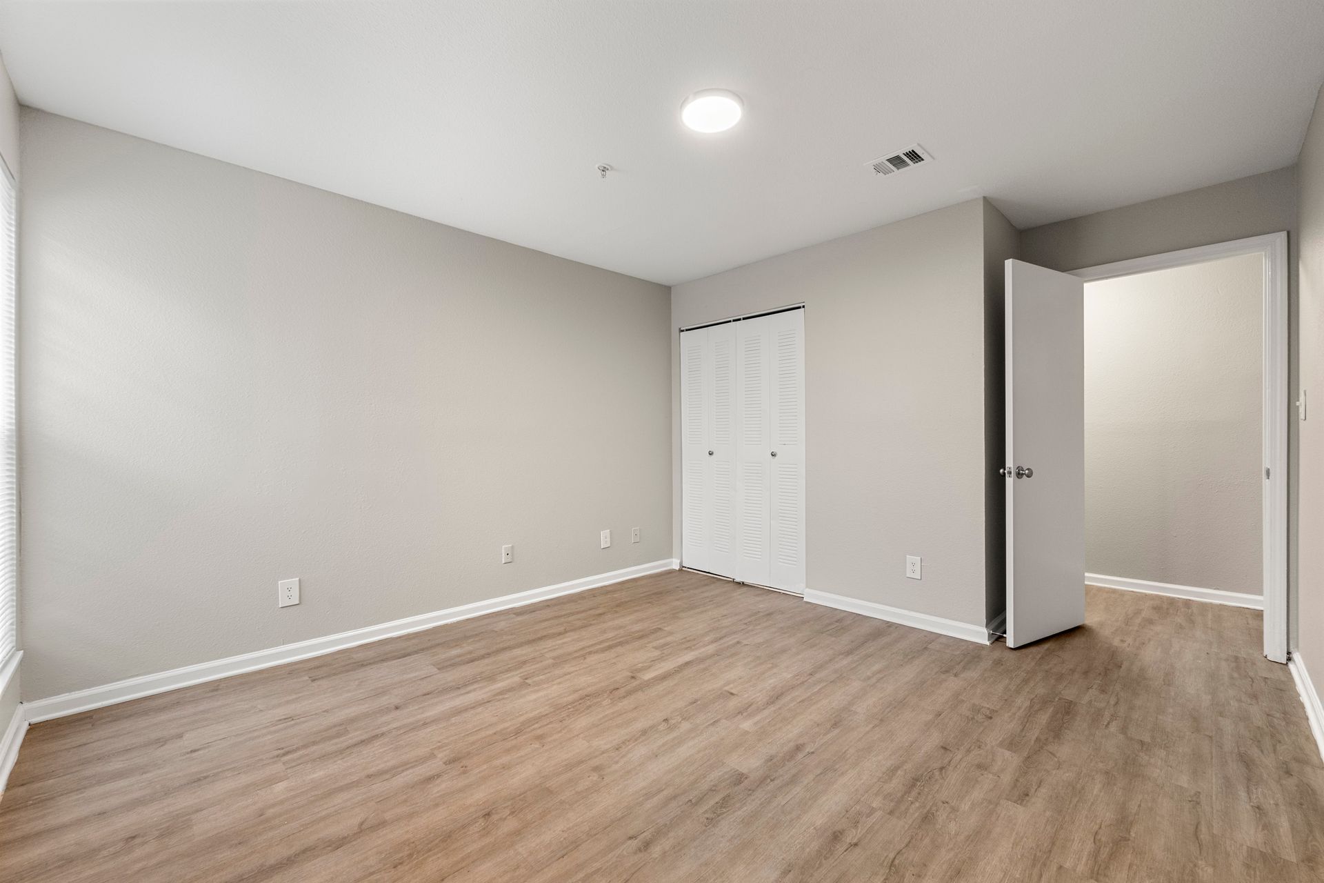 Empty beige bedroom with wood flooring, white closet doors, and an open door to a hallway
