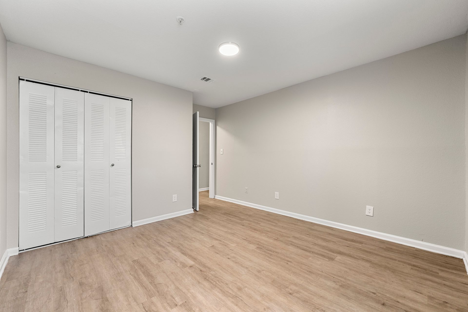 Empty beige bedroom with wood floors, white closet doors, and recessed ceiling light