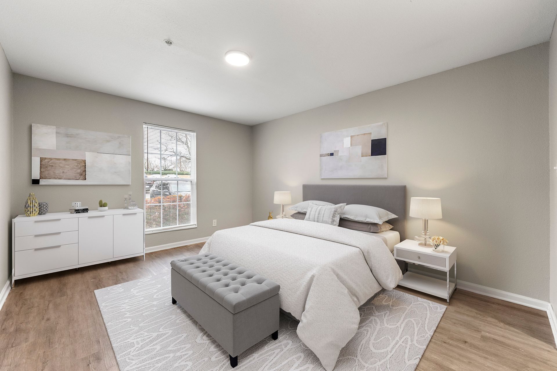 Bright bedroom with a gray bed, white dresser, nightstands, and a window on beige walls.
