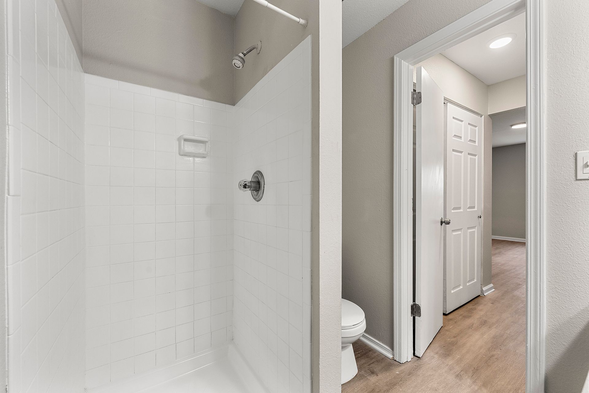 Bathroom with white-tiled shower, open door, and hallway with beige carpet beyond.
