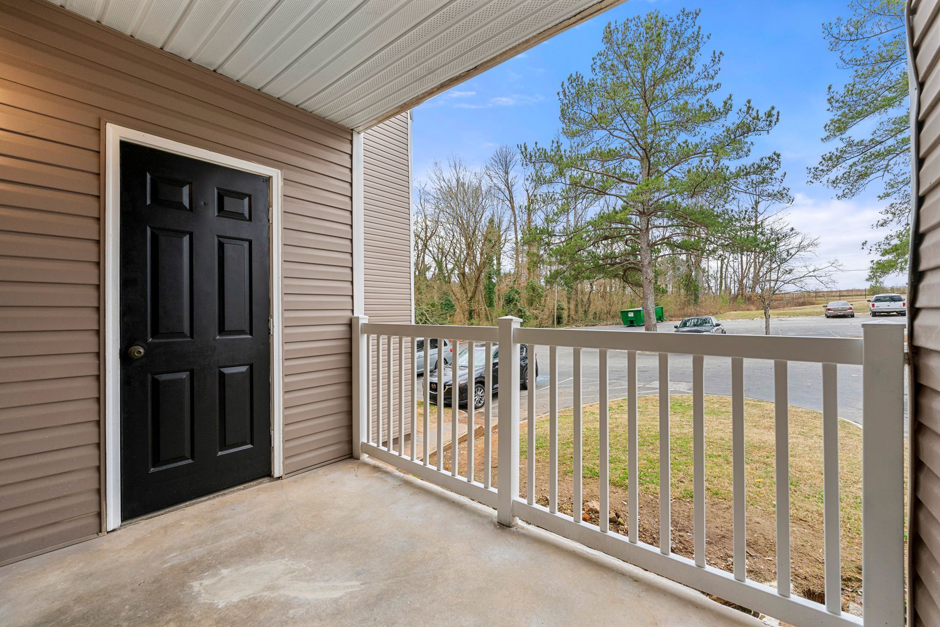 Apartment porch with a black door, white railing, and a view of trees and parked cars.