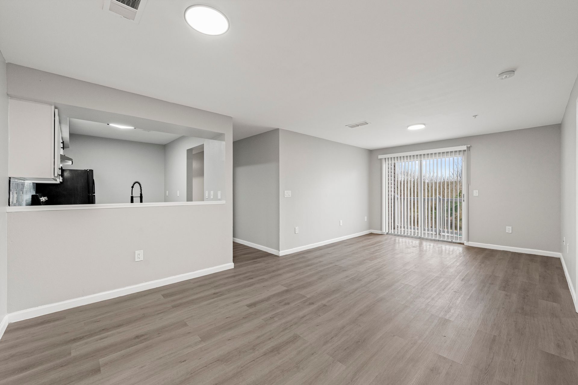 Empty open-plan living room with gray wood floors, white walls, and sliding glass doors to a patio.