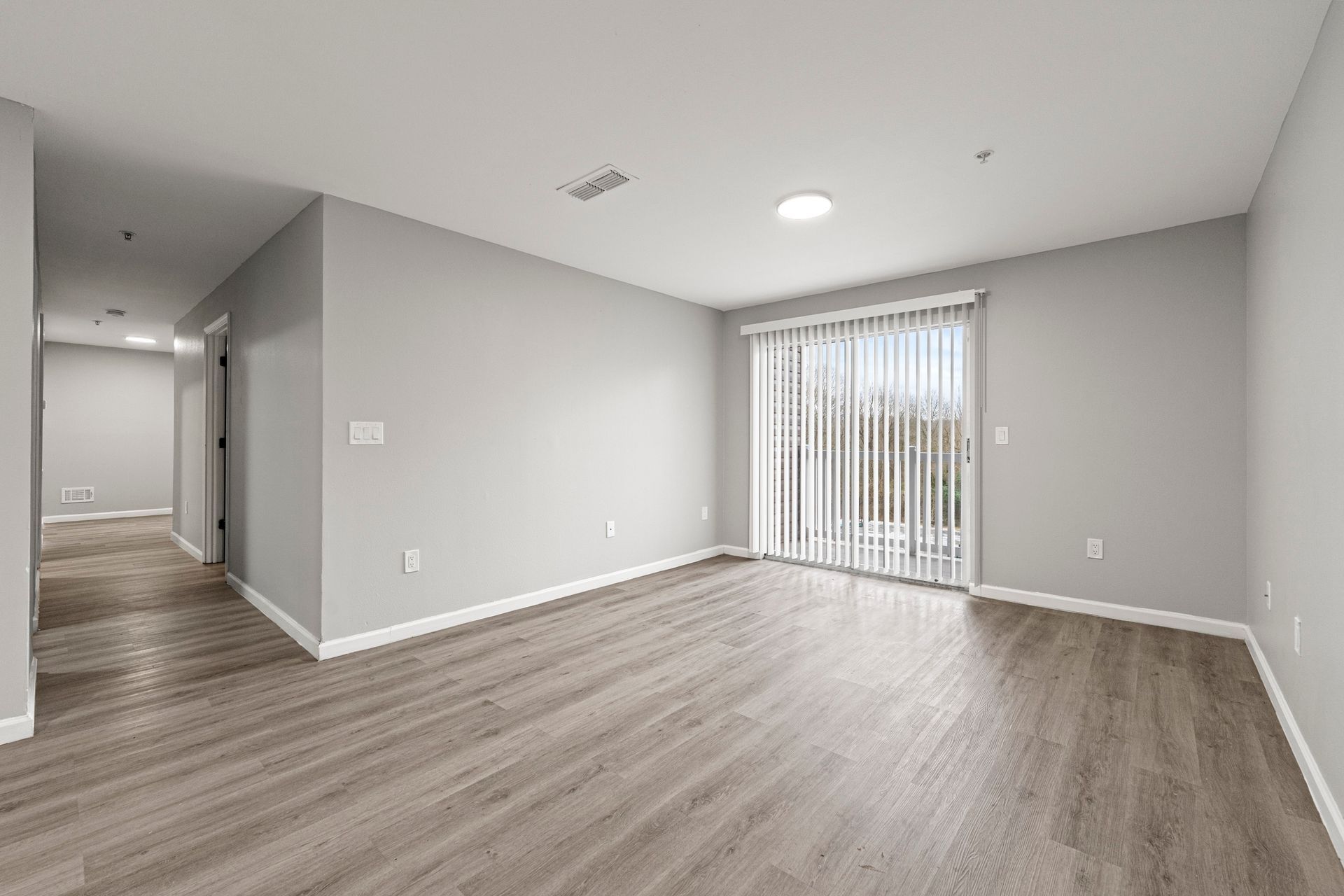 Empty modern living room with gray walls, light wood floors, and sliding glass doors with vertical blinds.