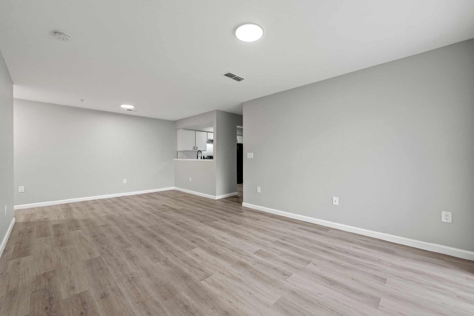 Empty gray living room with wood-look flooring and recessed ceiling lights, view toward a kitchen opening