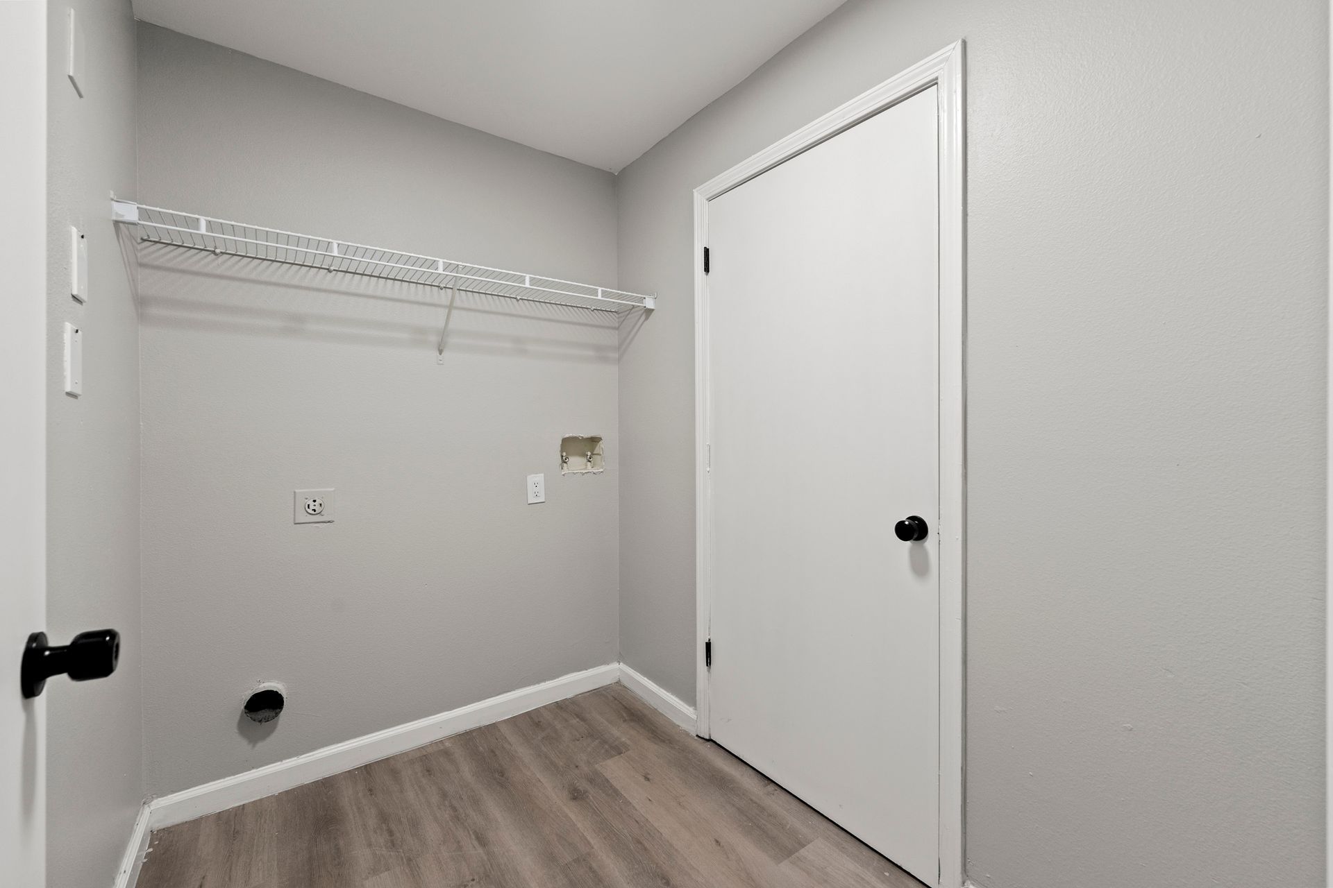 Empty laundry room with white walls, wire shelf, utility hookups, and a closed door.