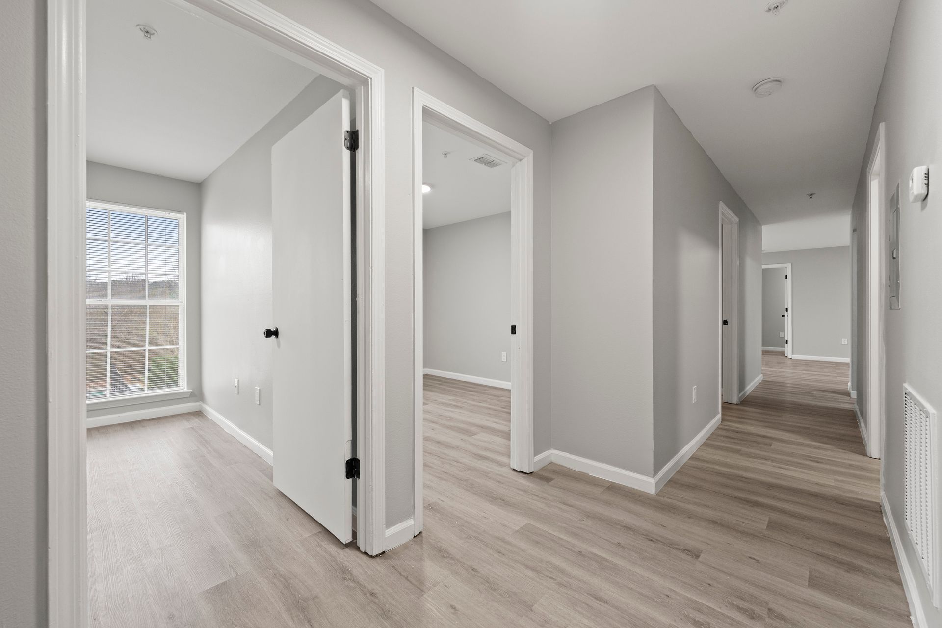 Bright empty hallway with white doors, gray walls, and wood flooring, leading to a sunlit room.